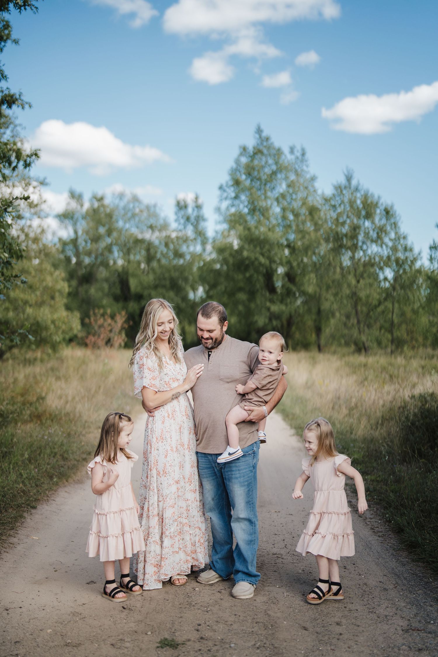 A group in coordinating pink and neutral outfits stands together on a dirt path surrounded by green trees on a sunny day.