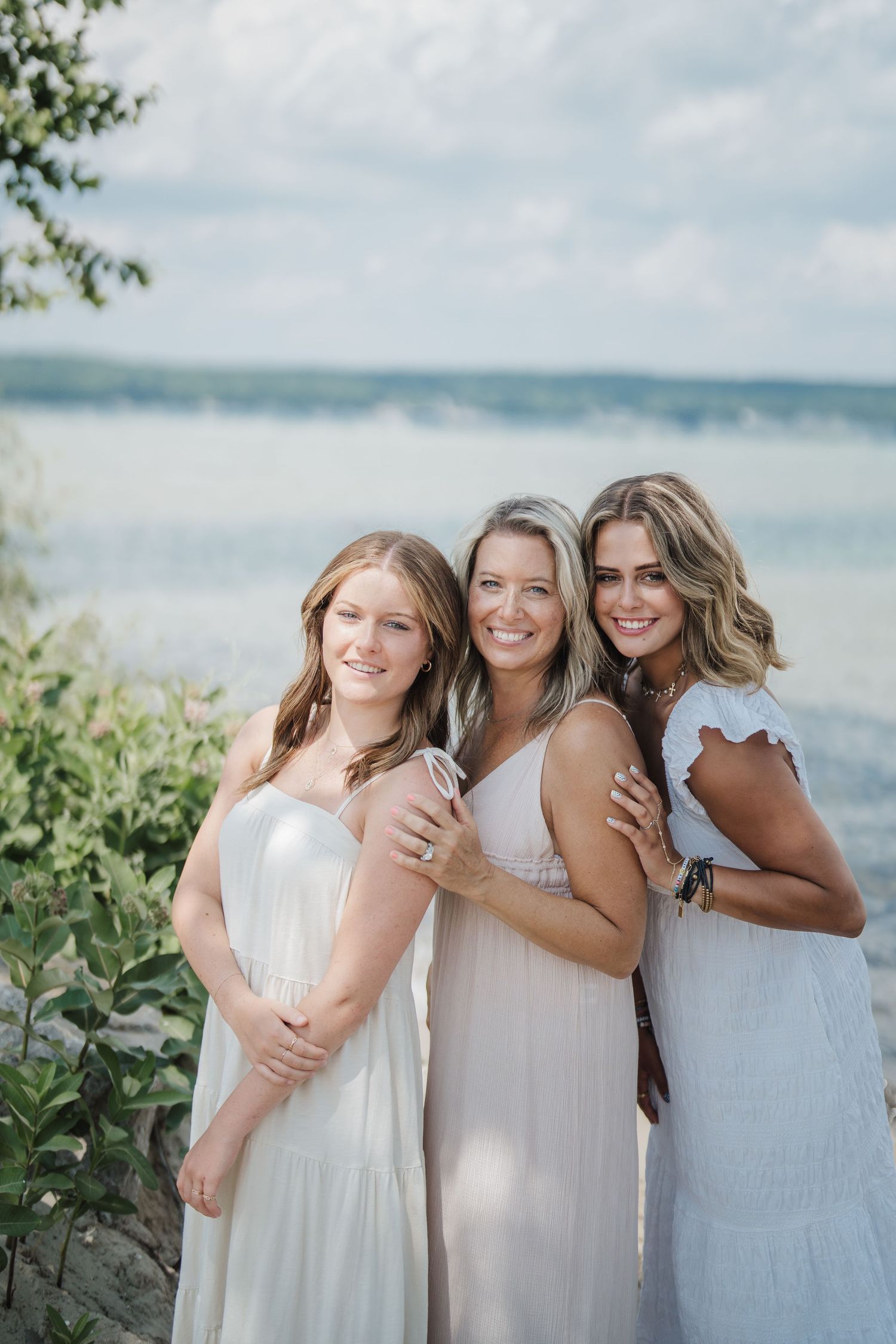 Three women in white summer dresses pose together on a beach with a scenic waterfront backdrop.