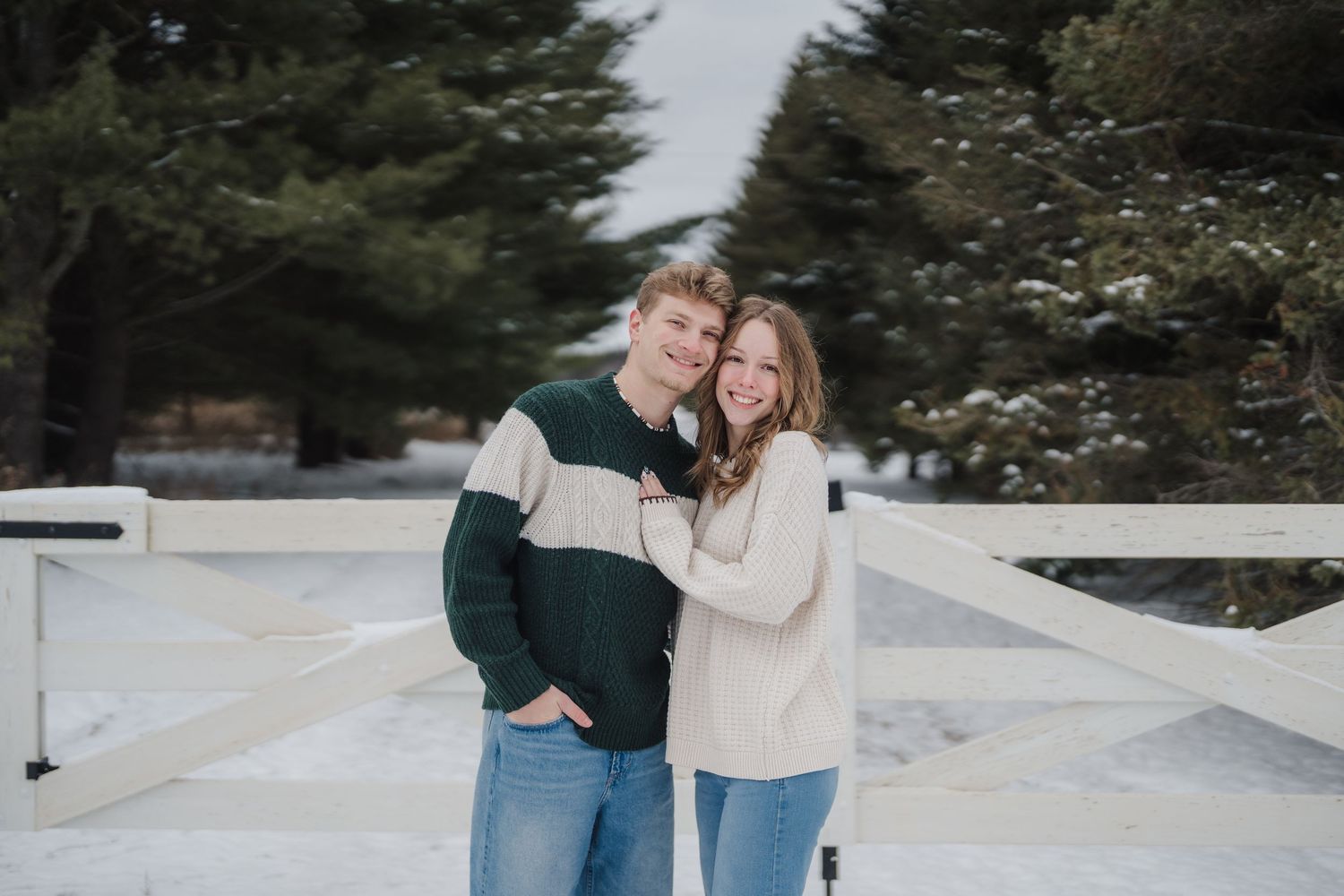 A couple in cozy sweaters embraces while standing in front of a white fence on a winter day.