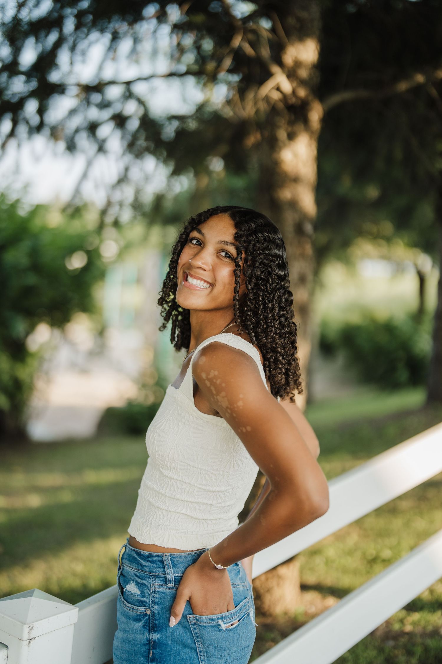 A casual summer portrait against a white fence featuring a white tank top and denim jeans in natural lighting.