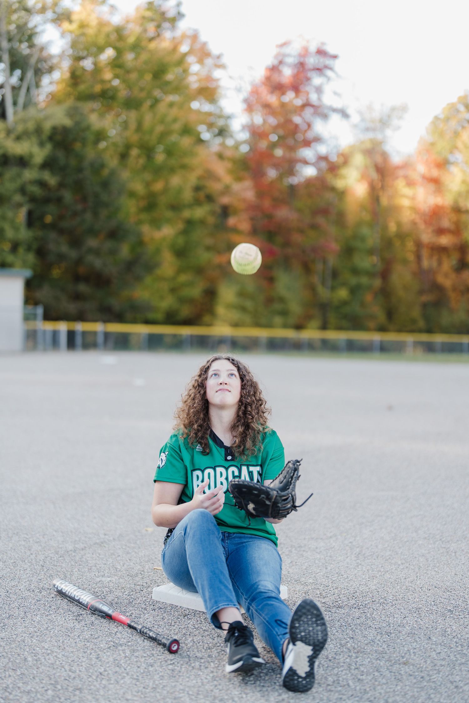 Person sitting on asphalt ground wearing jeans and green shirt with autumn trees in background during golden hour.