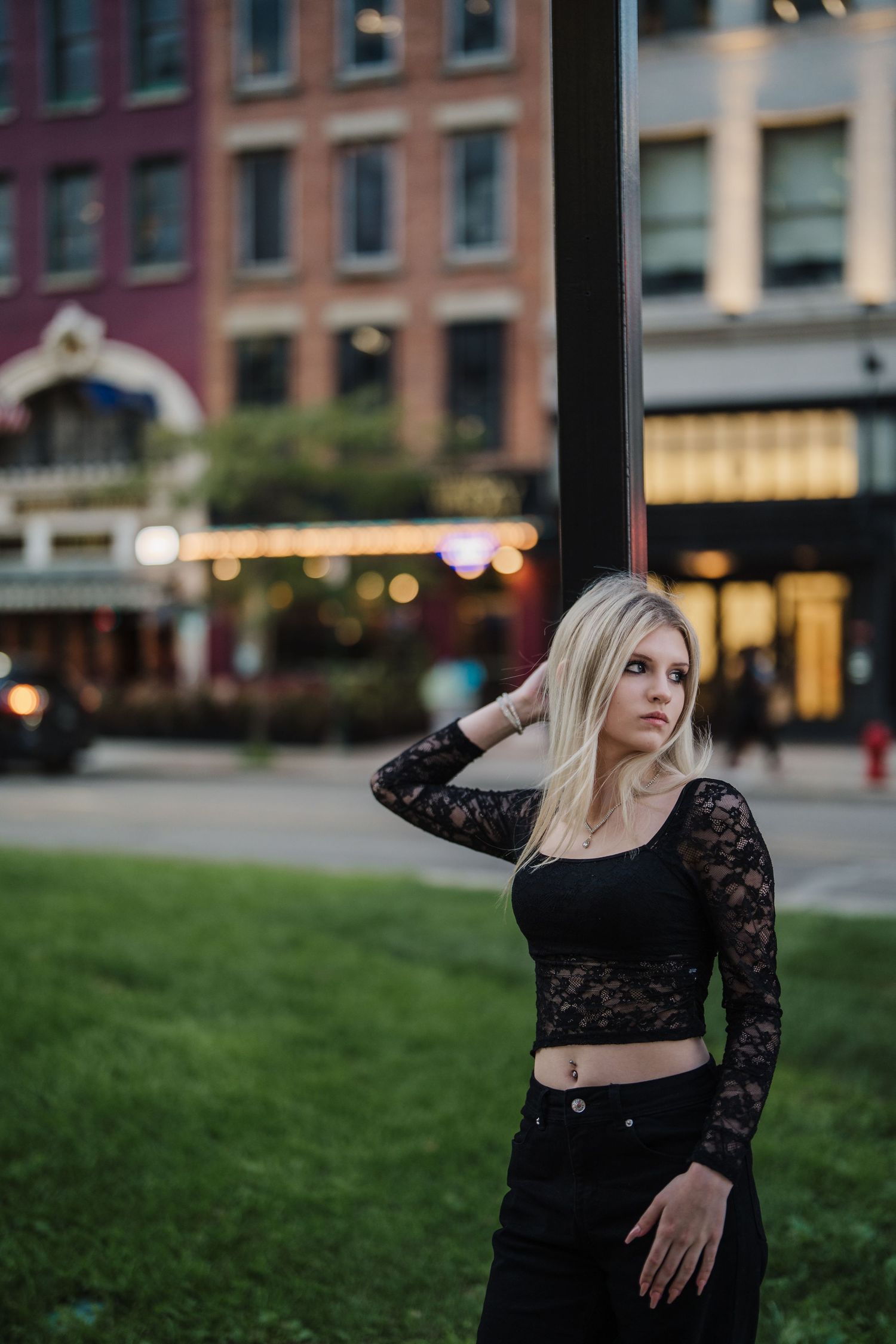 Fashion portrait of woman in black lace crop top posing against urban brick building backdrop at dusk.