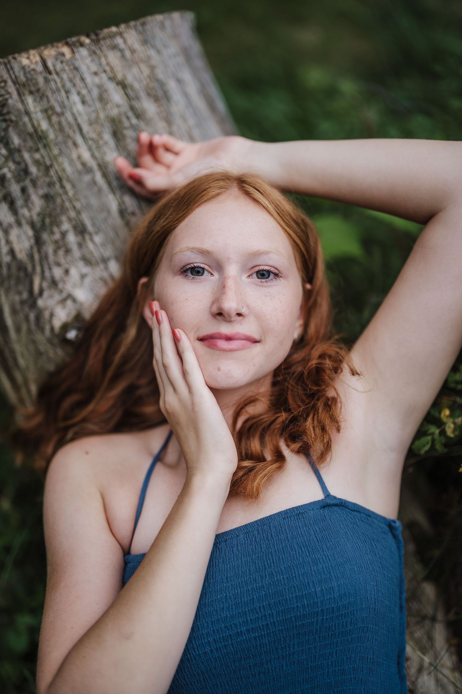 A portrait of someone with red hair wearing a blue top while resting against a tree trunk outdoors.