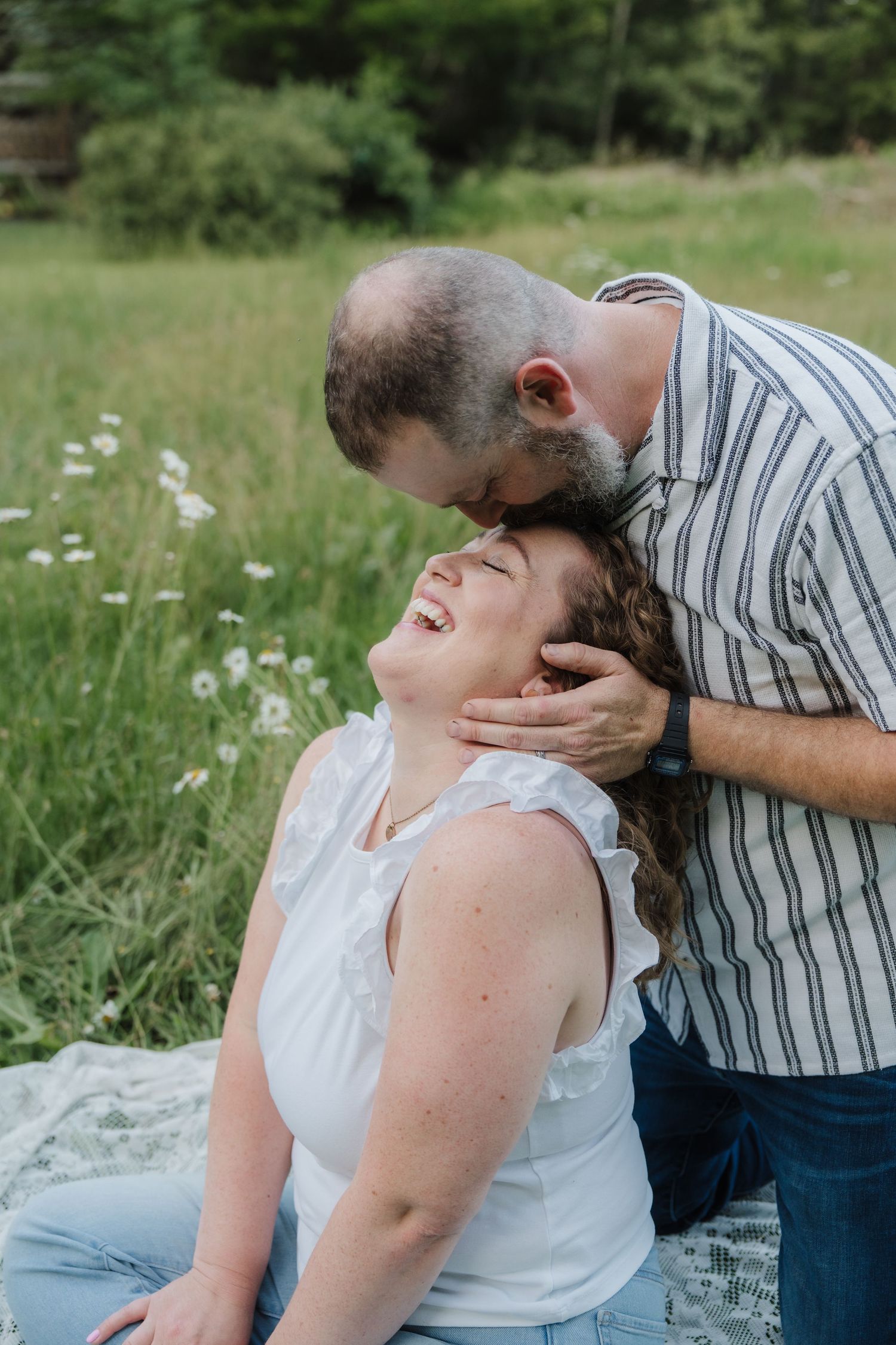 A man tenderly kisses a laughing woman's forehead while sitting in a sunny wildflower meadow.