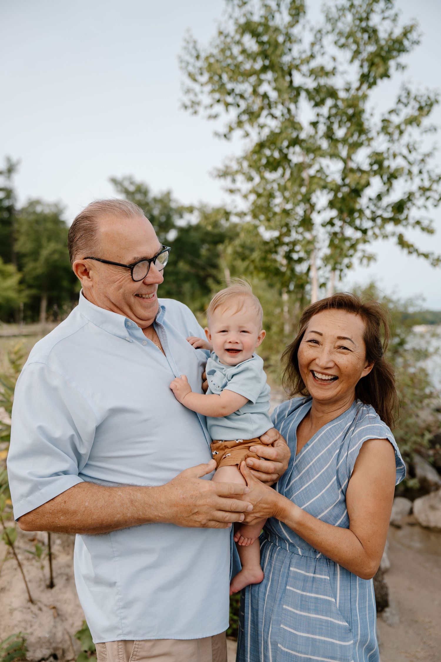 Smiling grandparents holding a happy baby outdoors near trees and a lake on a warm summer day.