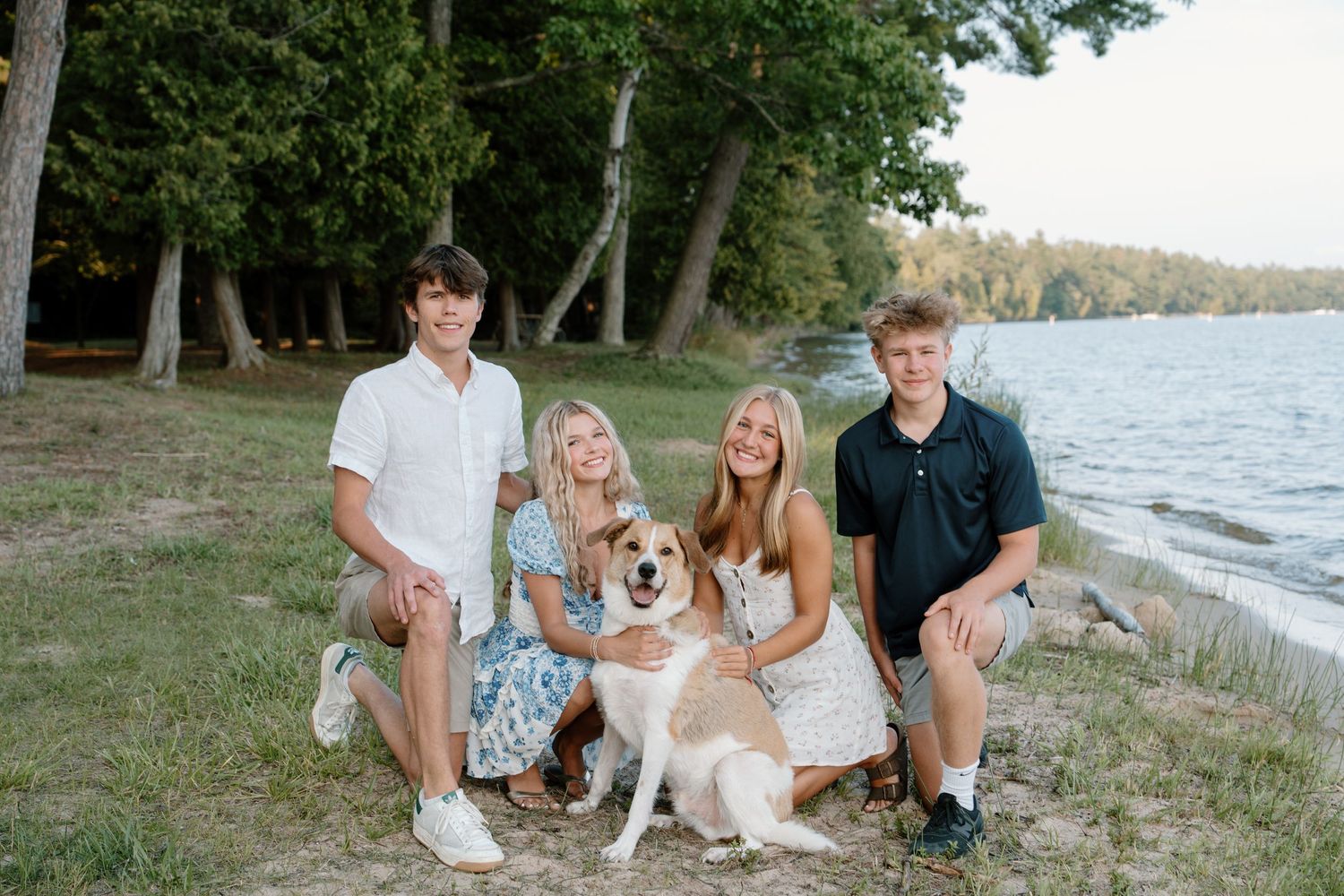 Family of four poses with their dog on a sandy lakeside beach surrounded by trees during golden hour.