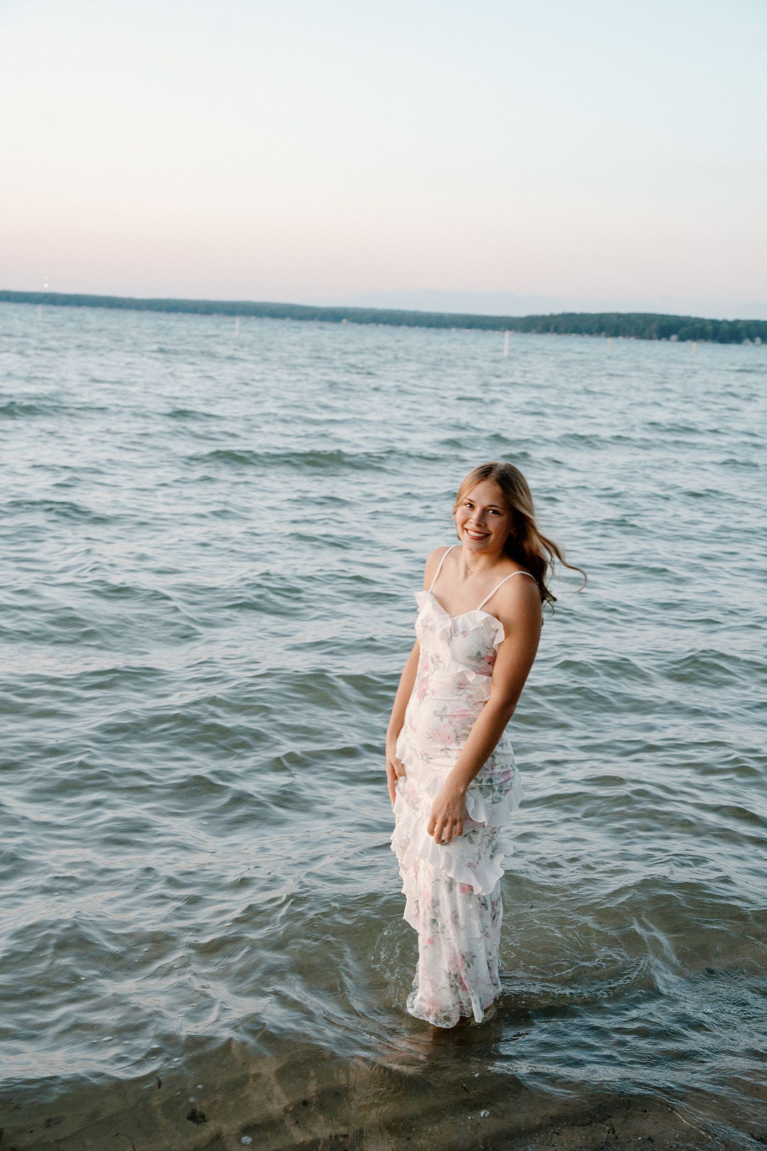 Young woman in floral dress standing in shallow lake water at sunset, smiling with wind in her hair.