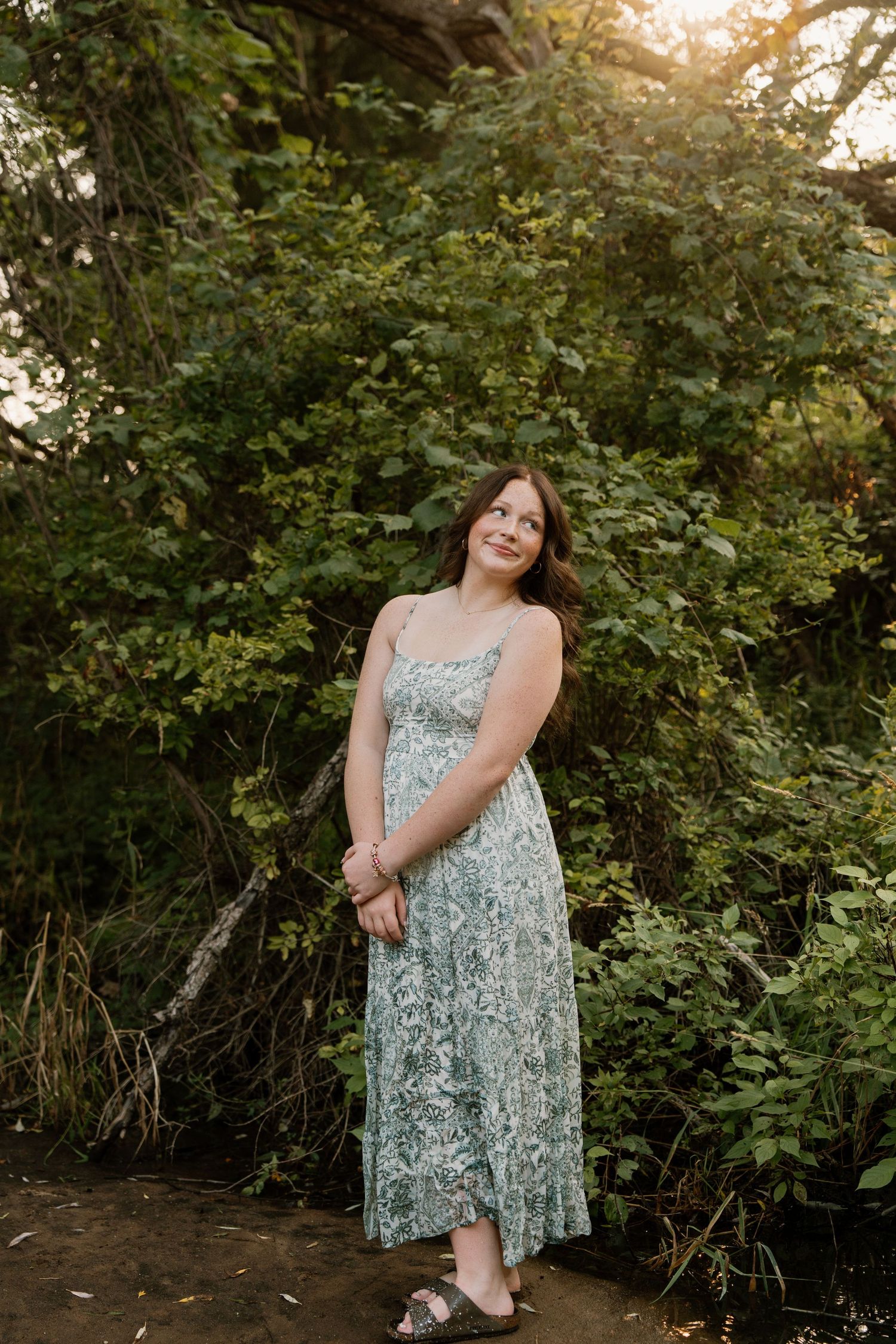 Young woman in floral maxi dress standing outdoors surrounded by lush green trees in golden hour light.