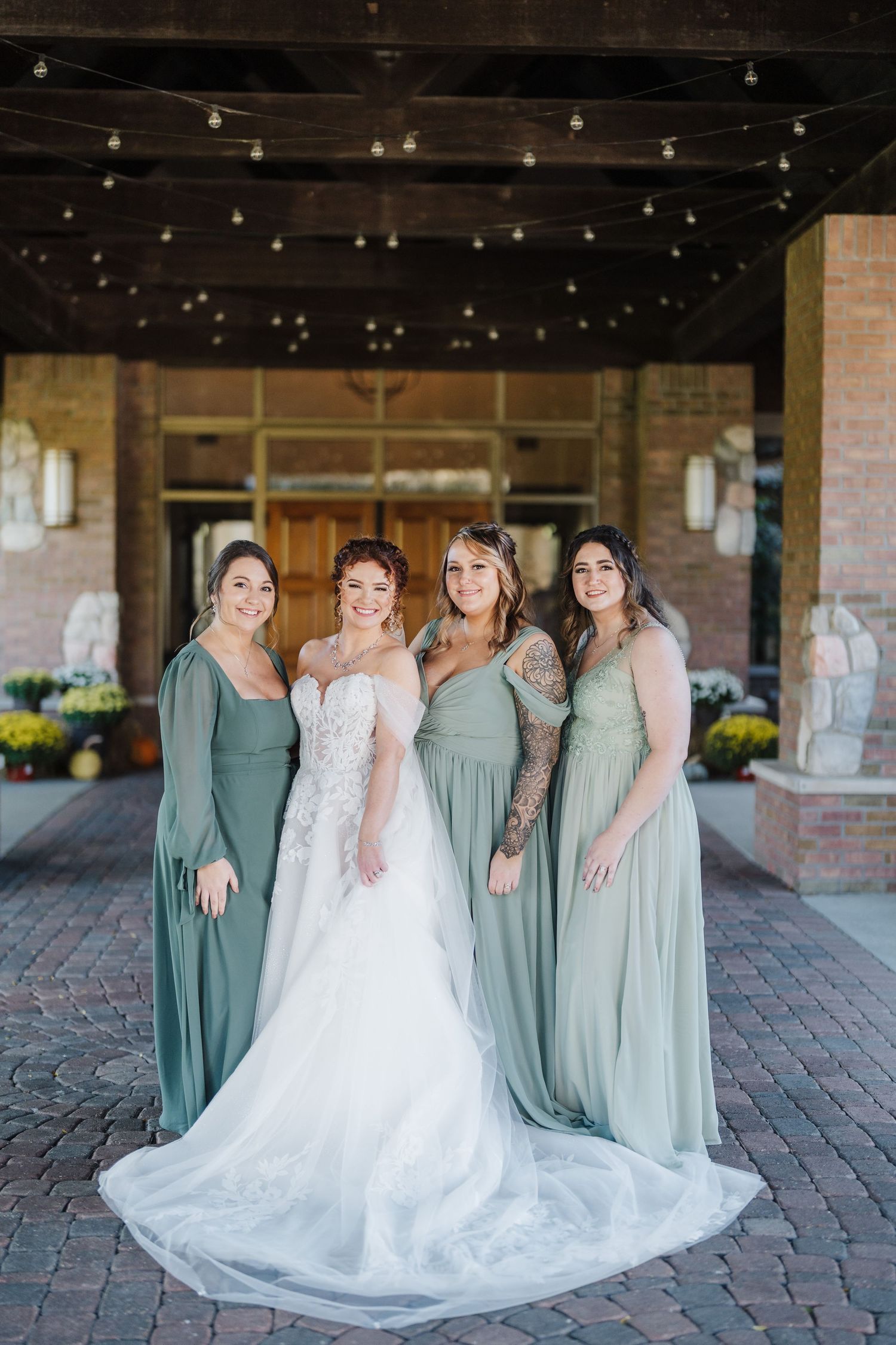 Bridal party poses in sage green bridesmaid dresses and white wedding gown under a covered brick patio entrance.