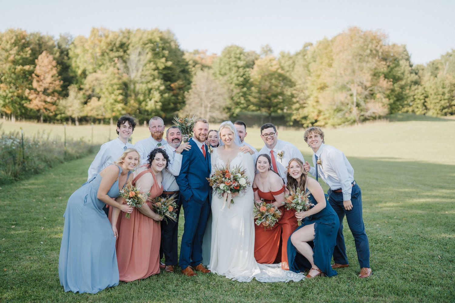 Wedding party members in coordinating blue and rust colored attire pose together on a grassy lawn at sunset.