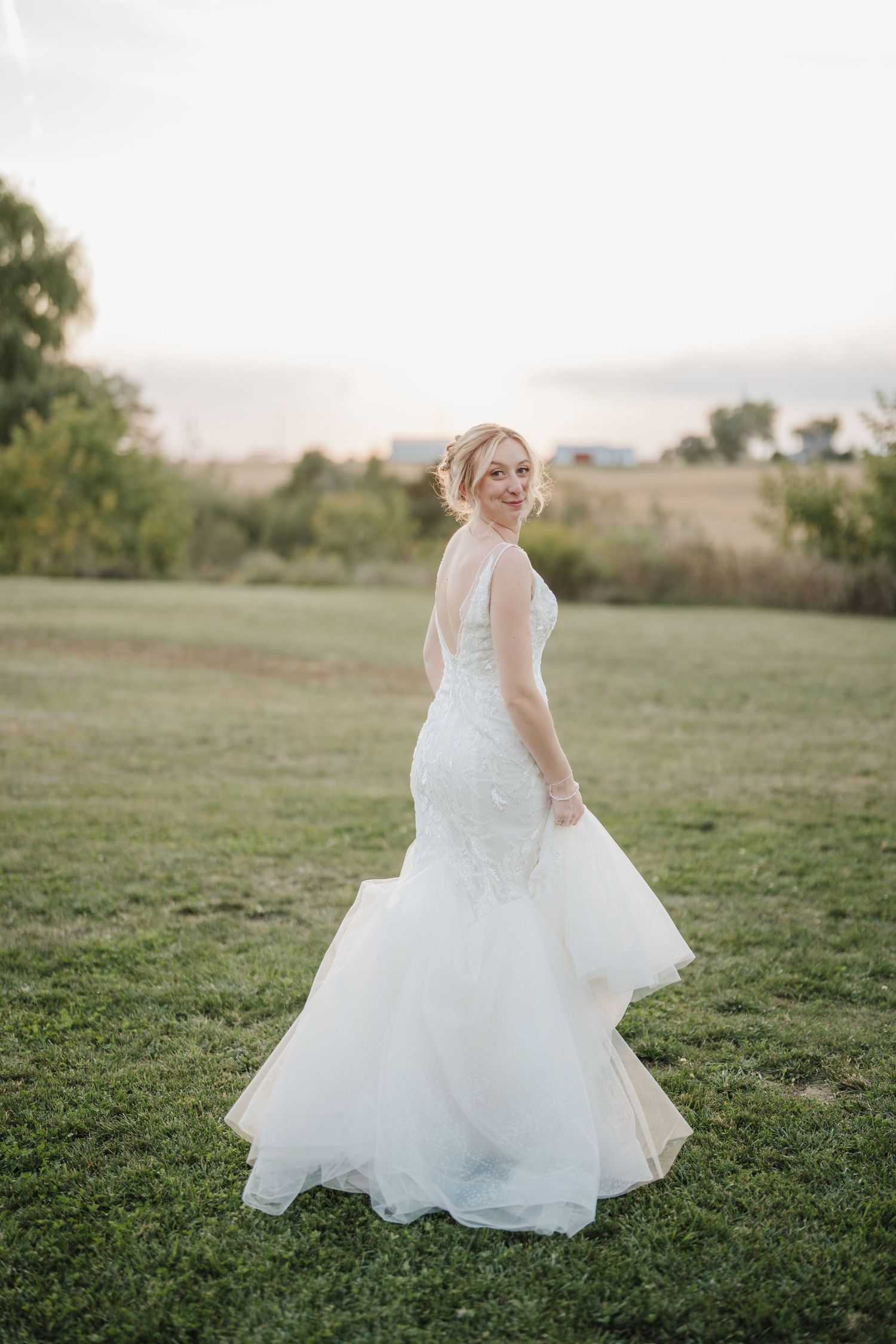 A bride in a flowing white mermaid-style wedding gown twirls gracefully on a grassy hillside at sunset.