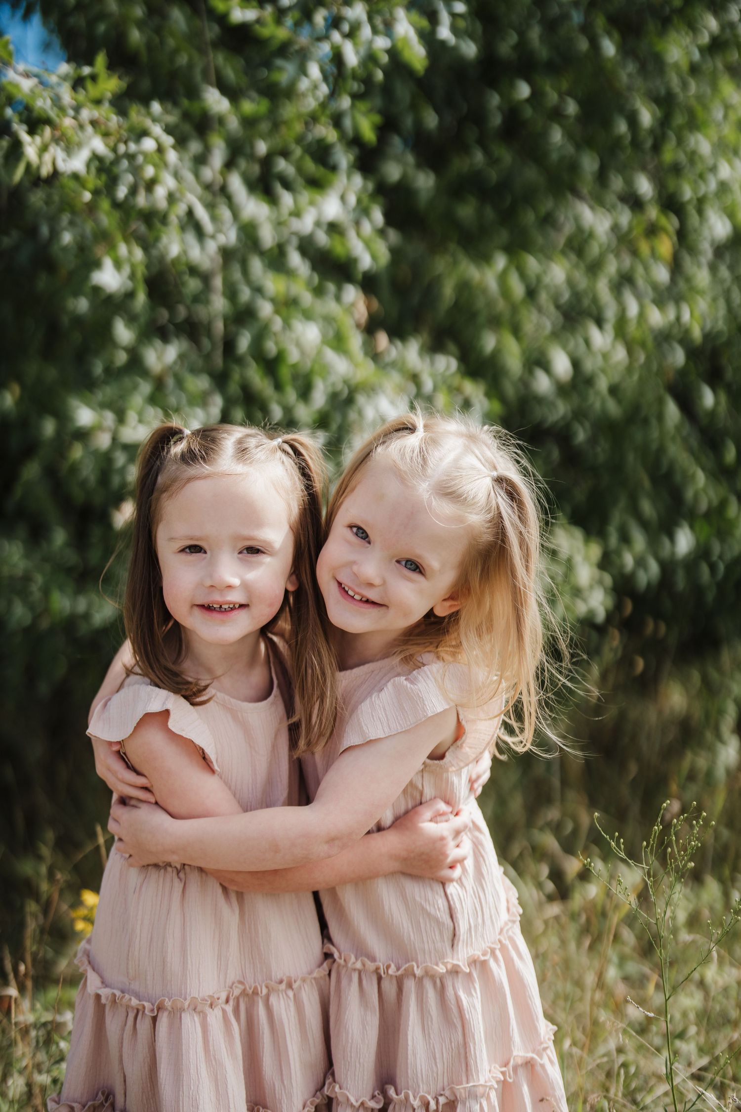 Two young children in matching pink dresses embrace while standing in a sunny outdoor setting with green foliage.