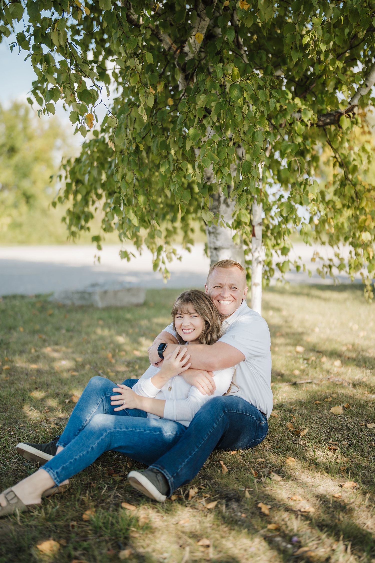 Two people sit together under a birch tree on a sunny day, sharing a warm embrace in casual denim outfits.