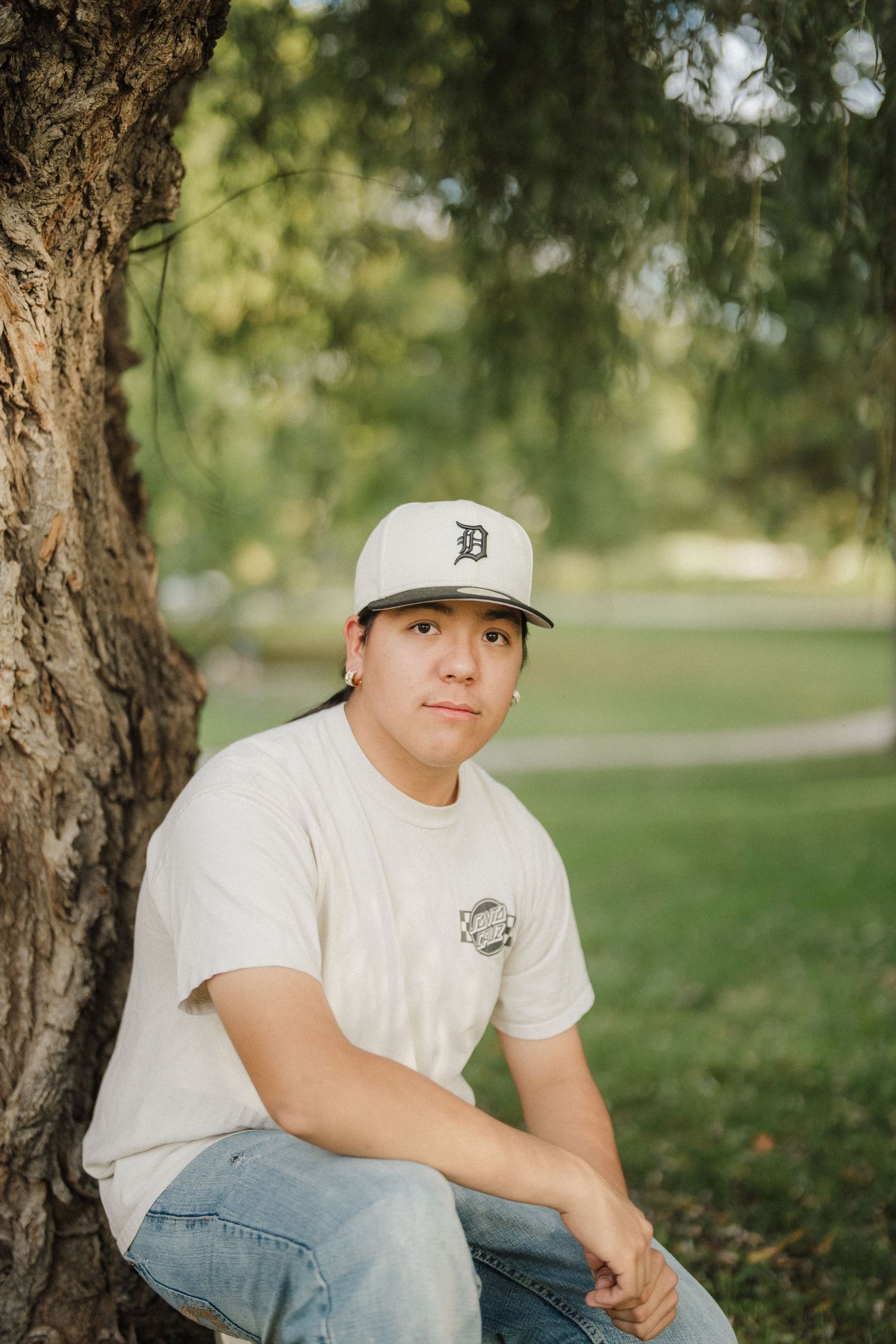 Person in white t-shirt and Detroit baseball cap sits under tree in outdoor setting.