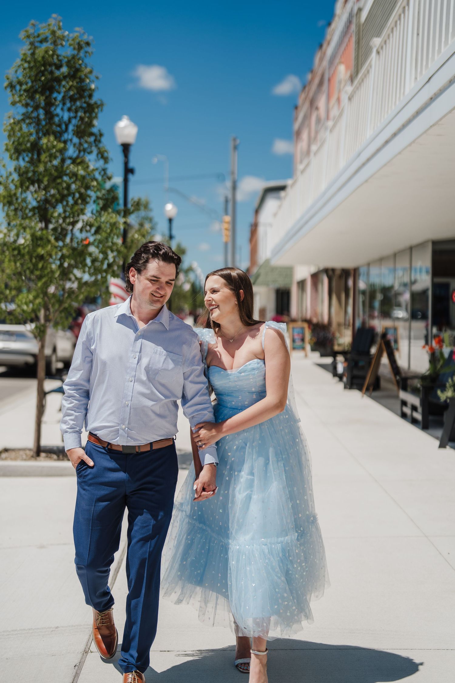 A couple shares joyful moments together on a sunny sidewalk in front of white storefronts.