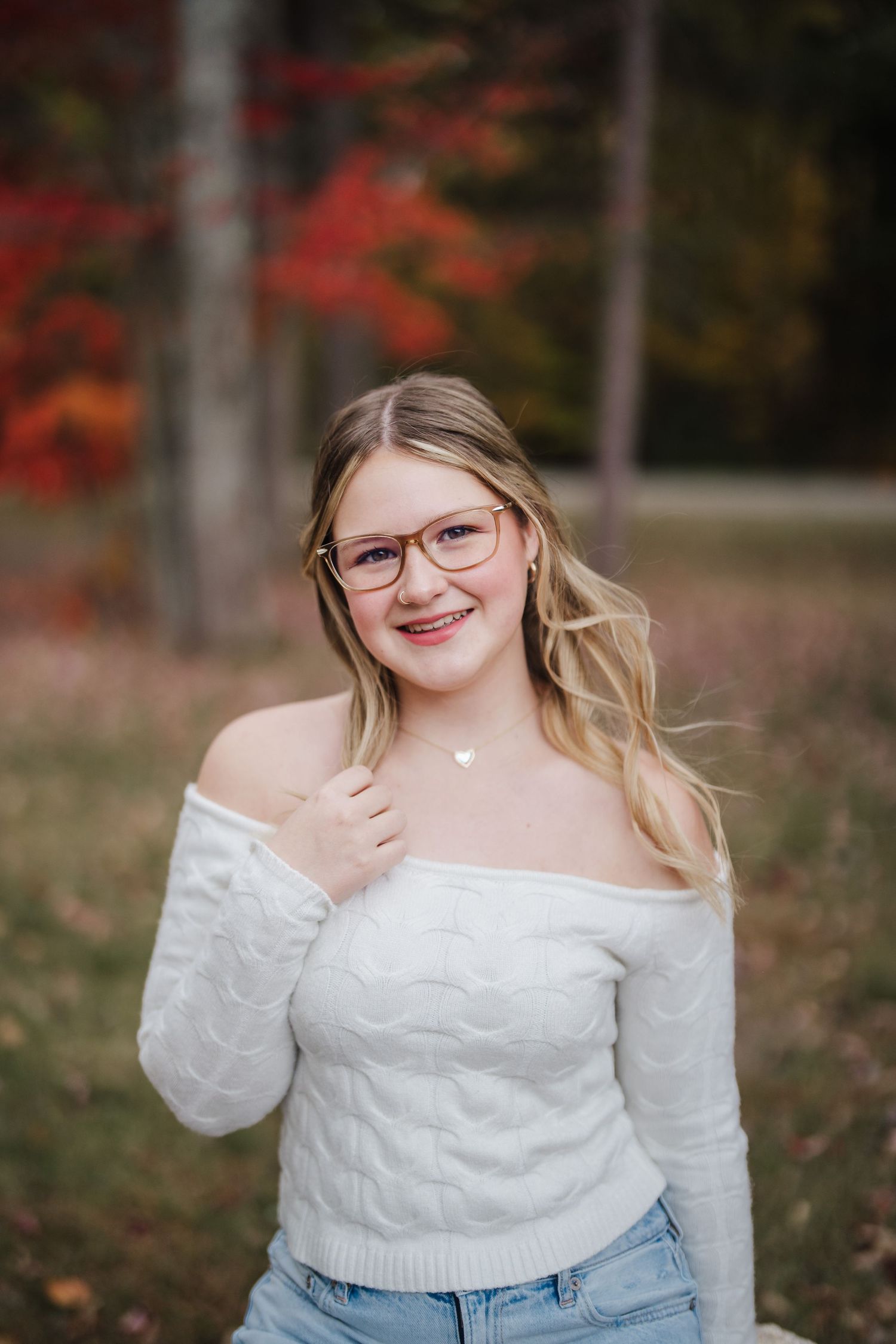 A person in a white sweater and jeans poses outdoors against a backdrop of autumn foliage.
