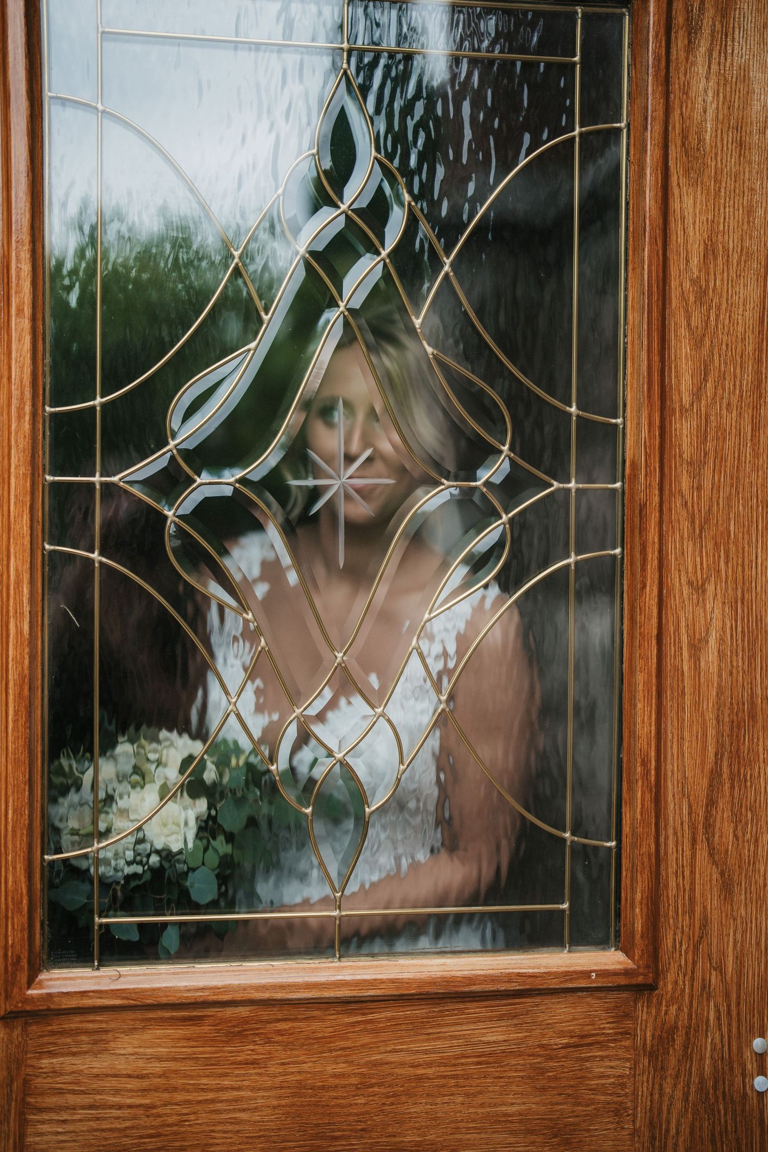 A close-up view through a decorative door window showing a bride holding a bouquet.