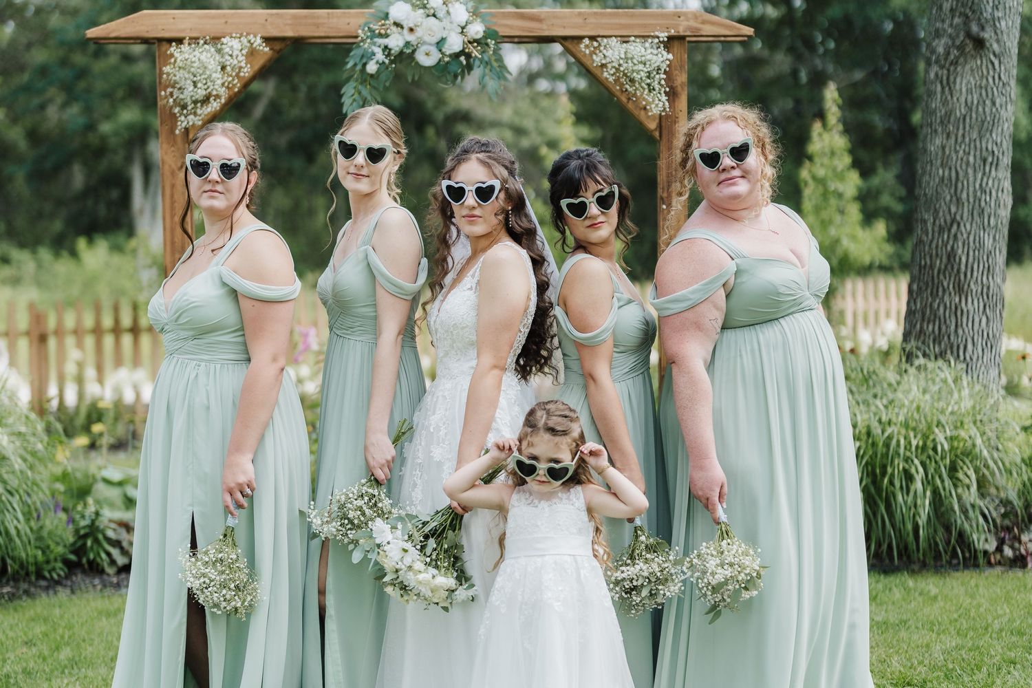 Bridesmaids and flower girl in mint green dresses pose wearing sunglasses in a garden setting.