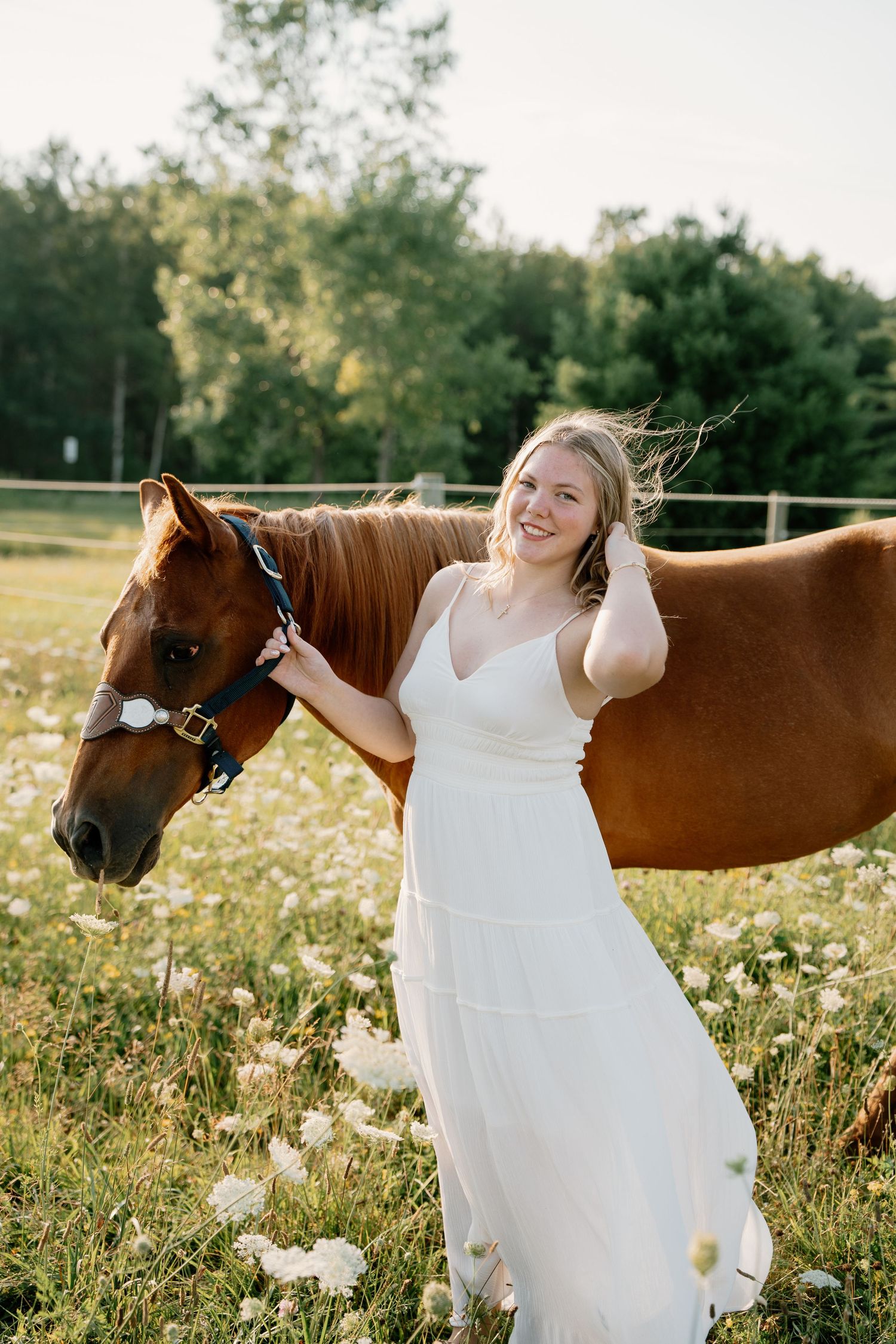 Smiling woman in white dress holding a chestnut horse in a sunny field of wildflowers.