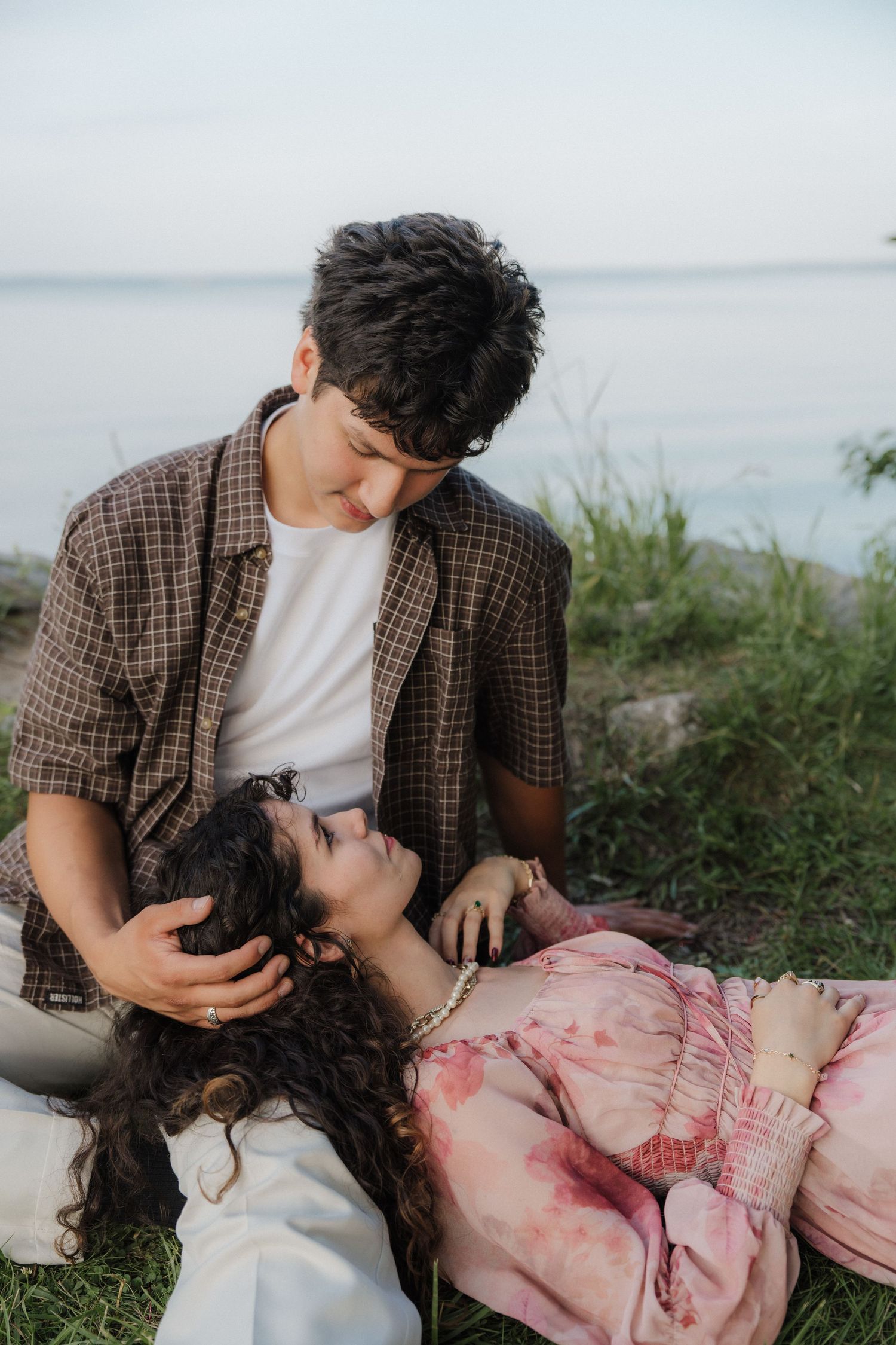 A young couple shares a tender moment by a lakeside, with the woman lying on grass looking up at the man.