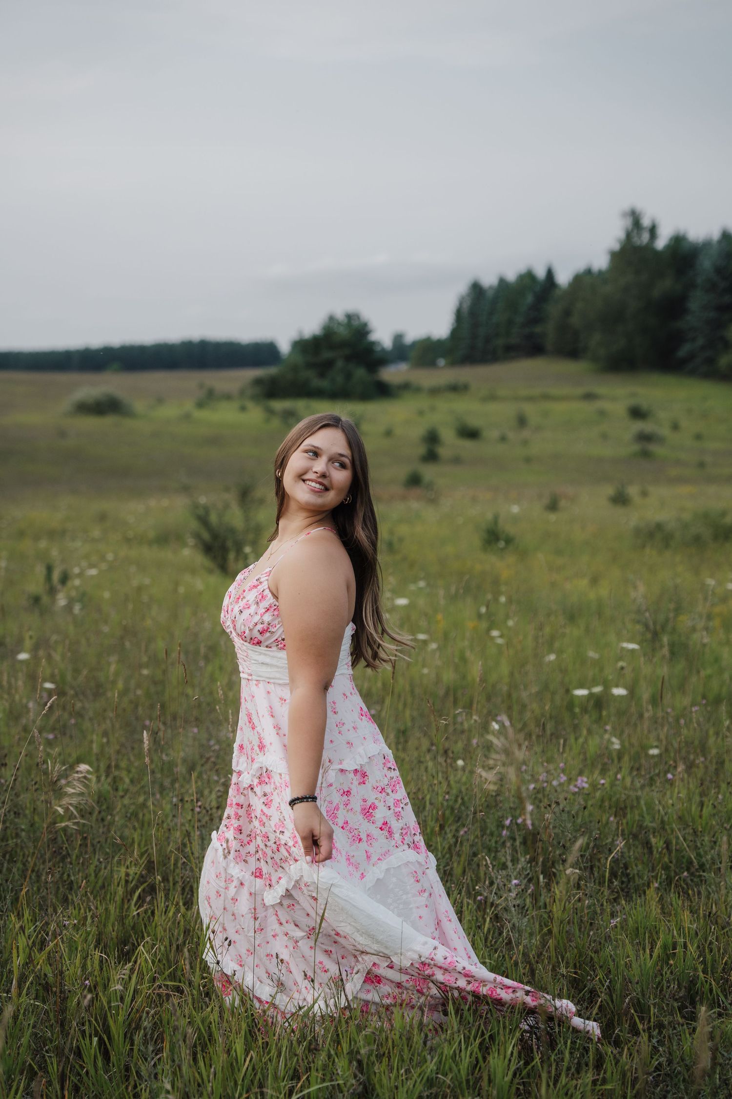 Smiling young woman in a floral pink dress standing in a green meadow with trees in the background.