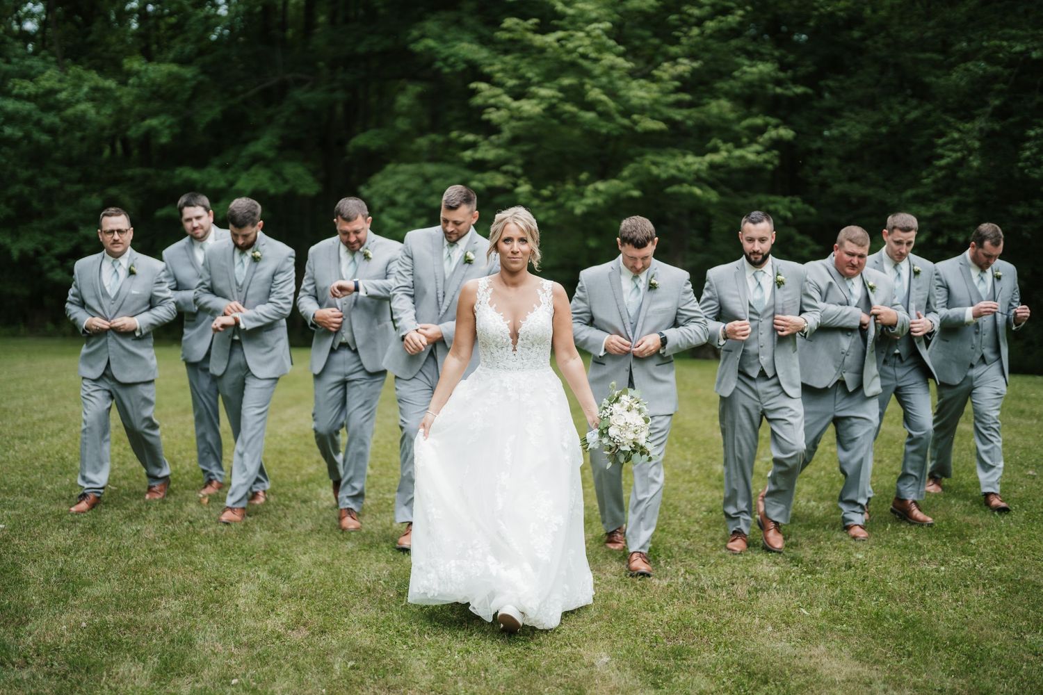 Wedding party in mint and gray attire poses playfully on grassy lawn during outdoor celebration.