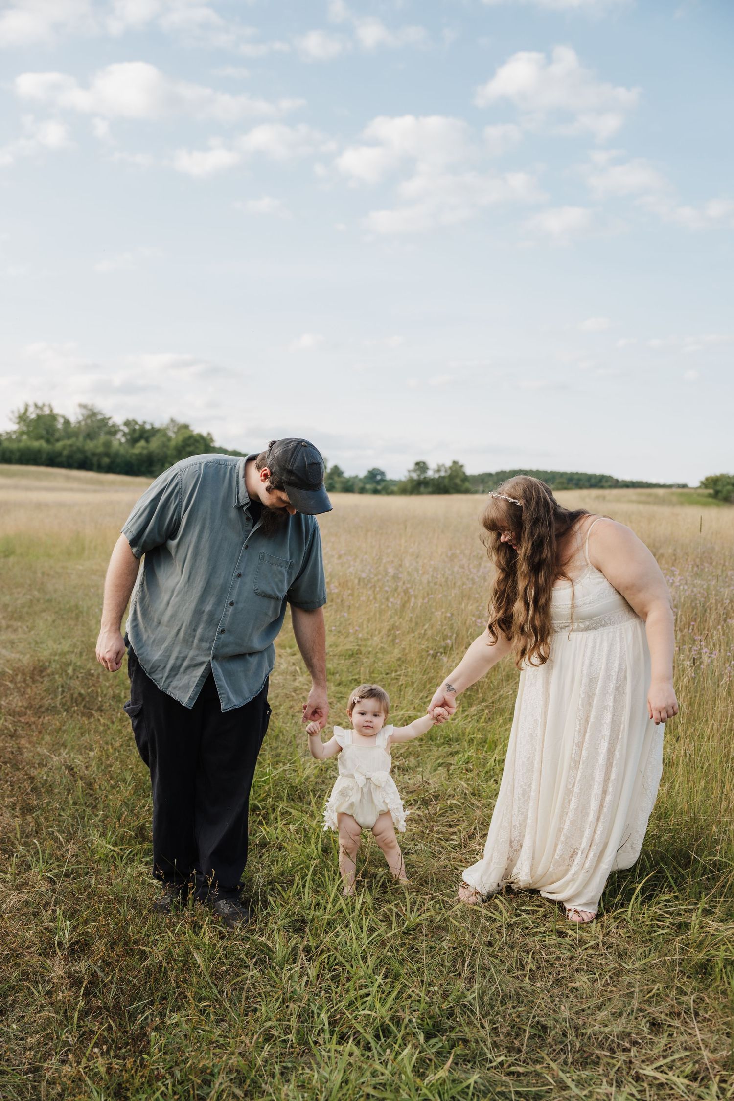 A couple holds their baby's hands while walking in a golden field under a partly cloudy sky.