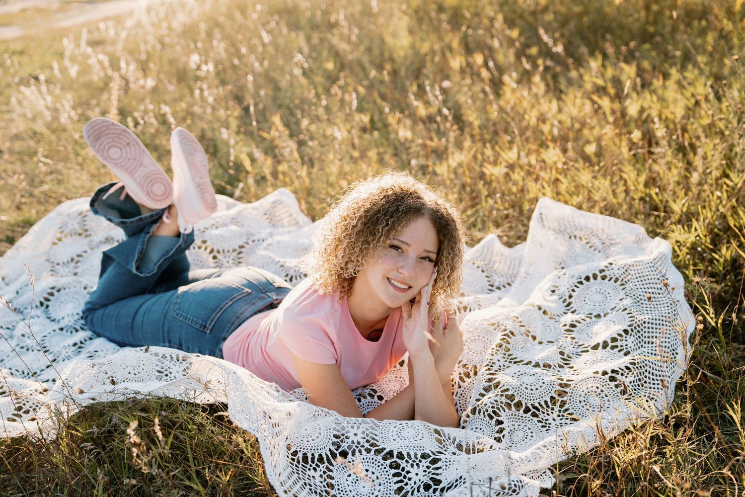 A young girl with curly hair lies on a lace blanket in a sunny field, smiling warmly at the camera.