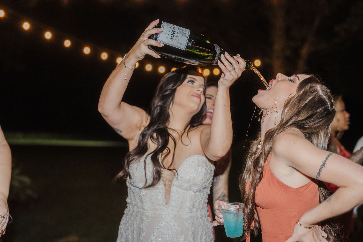 Wedding party members raise champagne glasses in a celebratory toast during an outdoor evening reception.