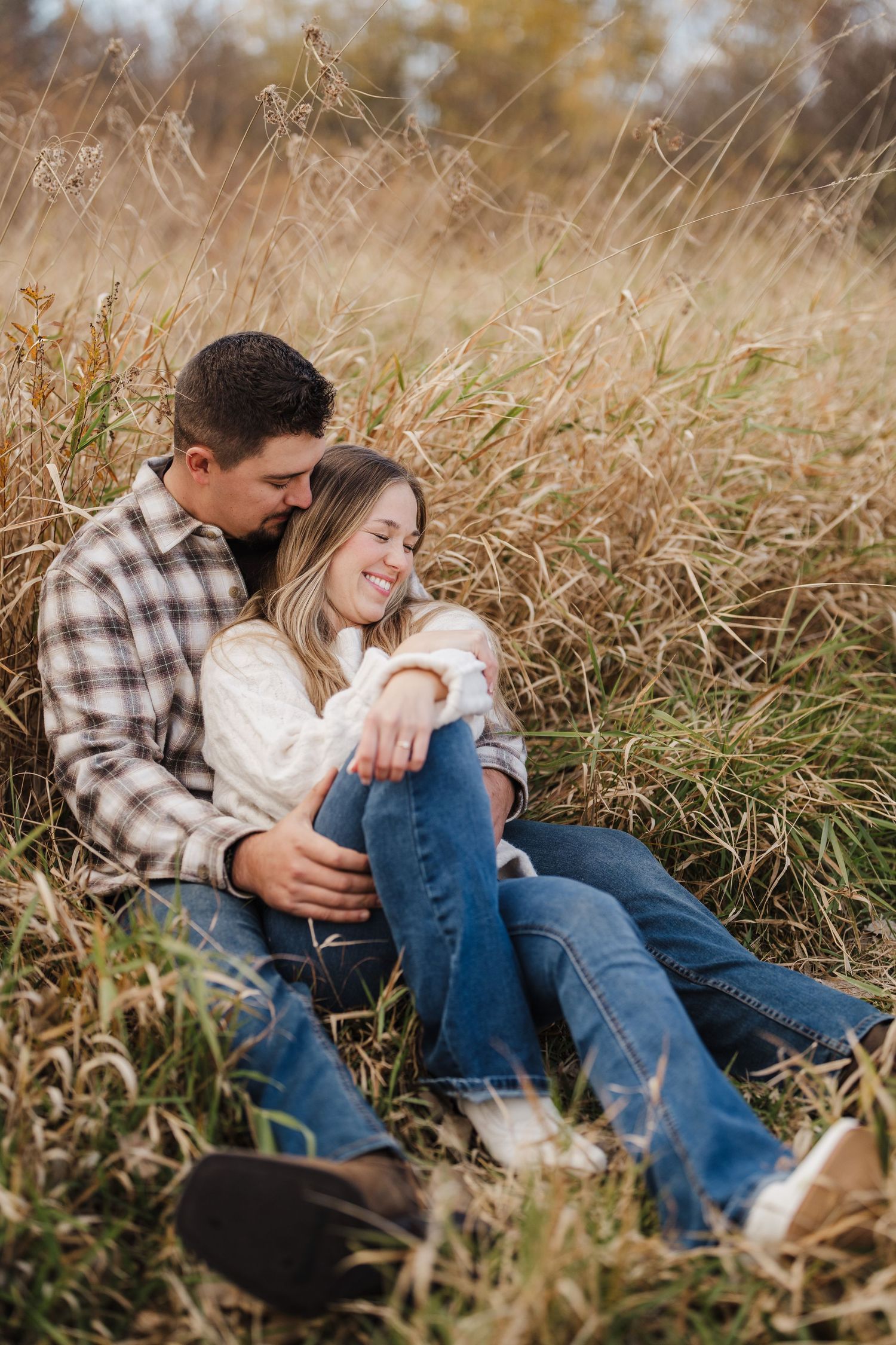 A couple sits together intimately in tall autumn grass during an outdoor photoshoot.