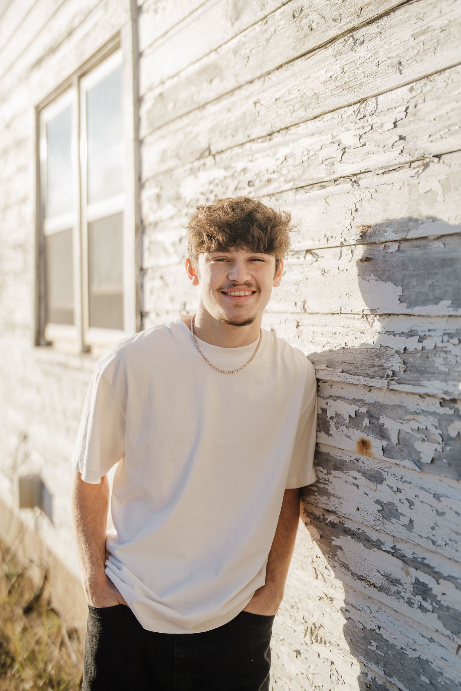 A person in a white t-shirt poses against a rustic white wall in warm sunlight.