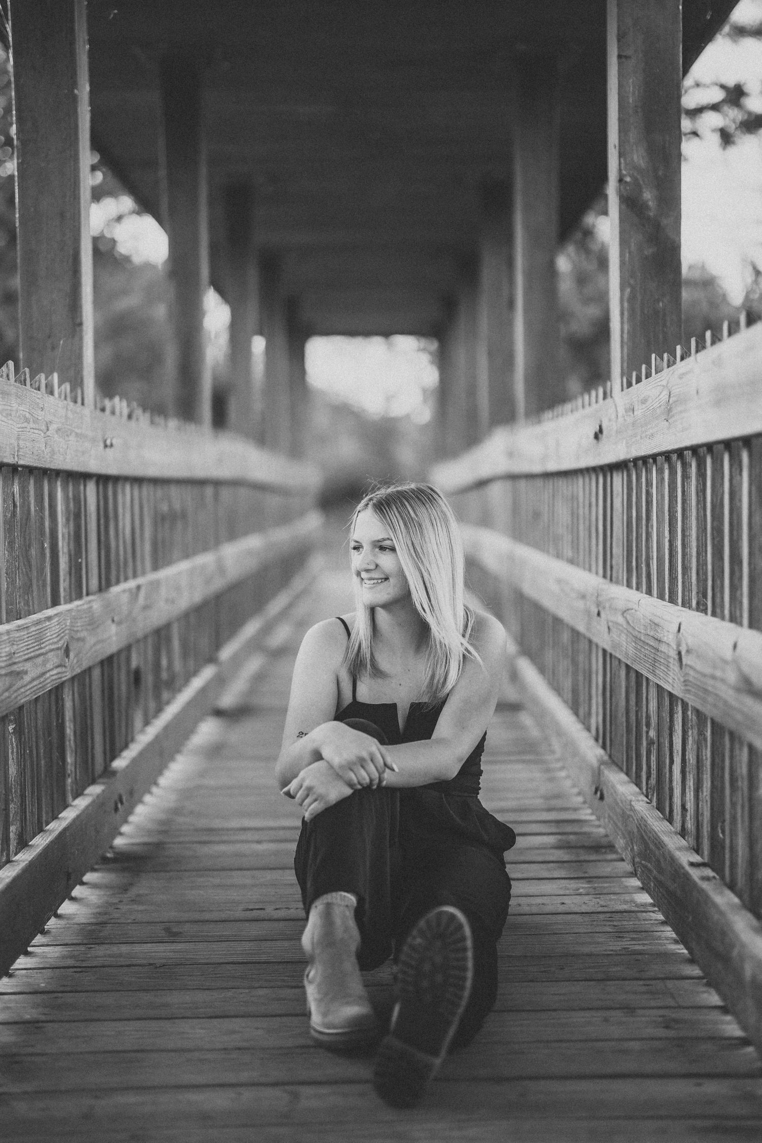 Black and white portrait of someone sitting casually on a wooden bridge walkway with covered roof overhead.