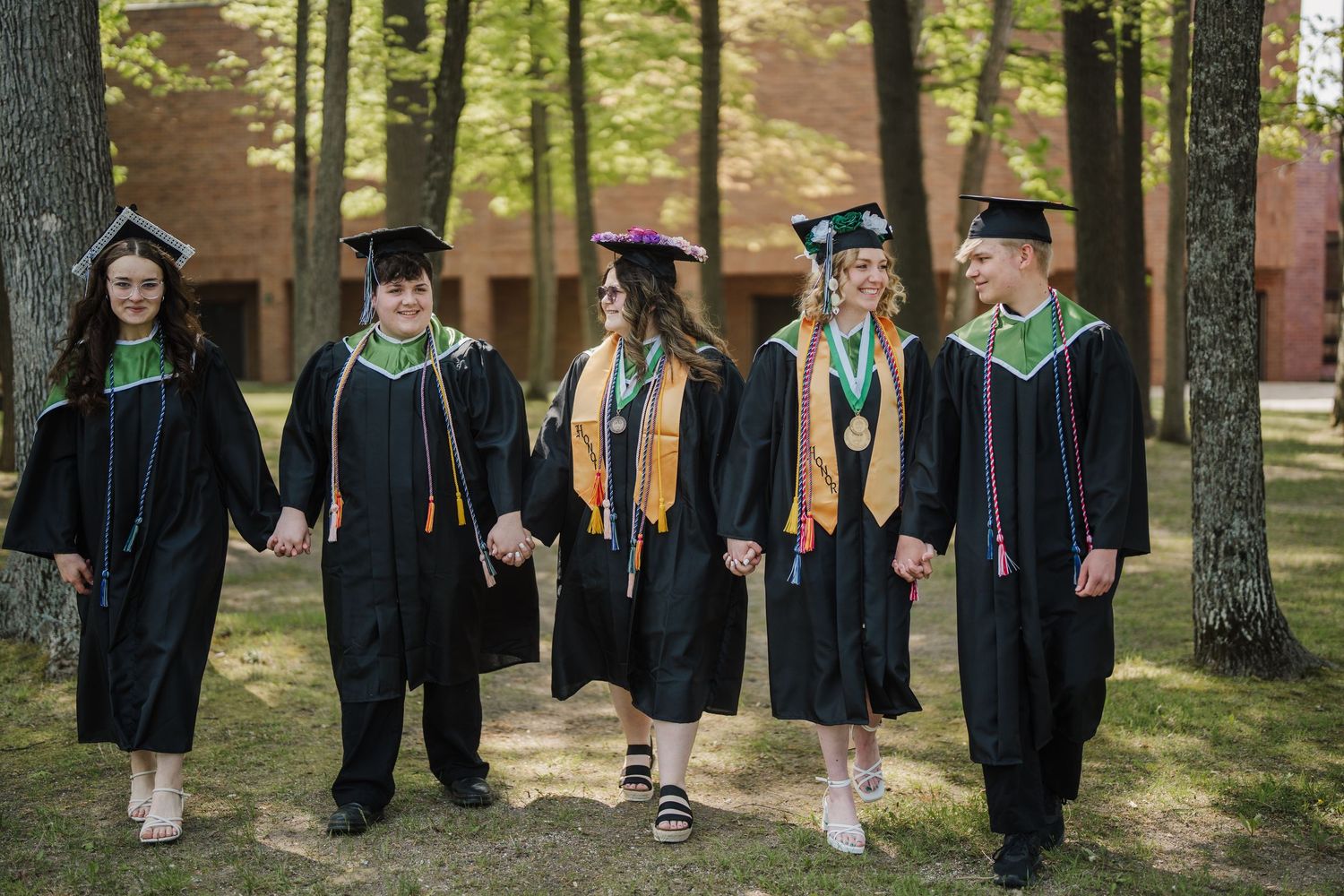 A group of graduates in black caps and gowns pose together outdoors on a sunny day near trees and brick buildings.
