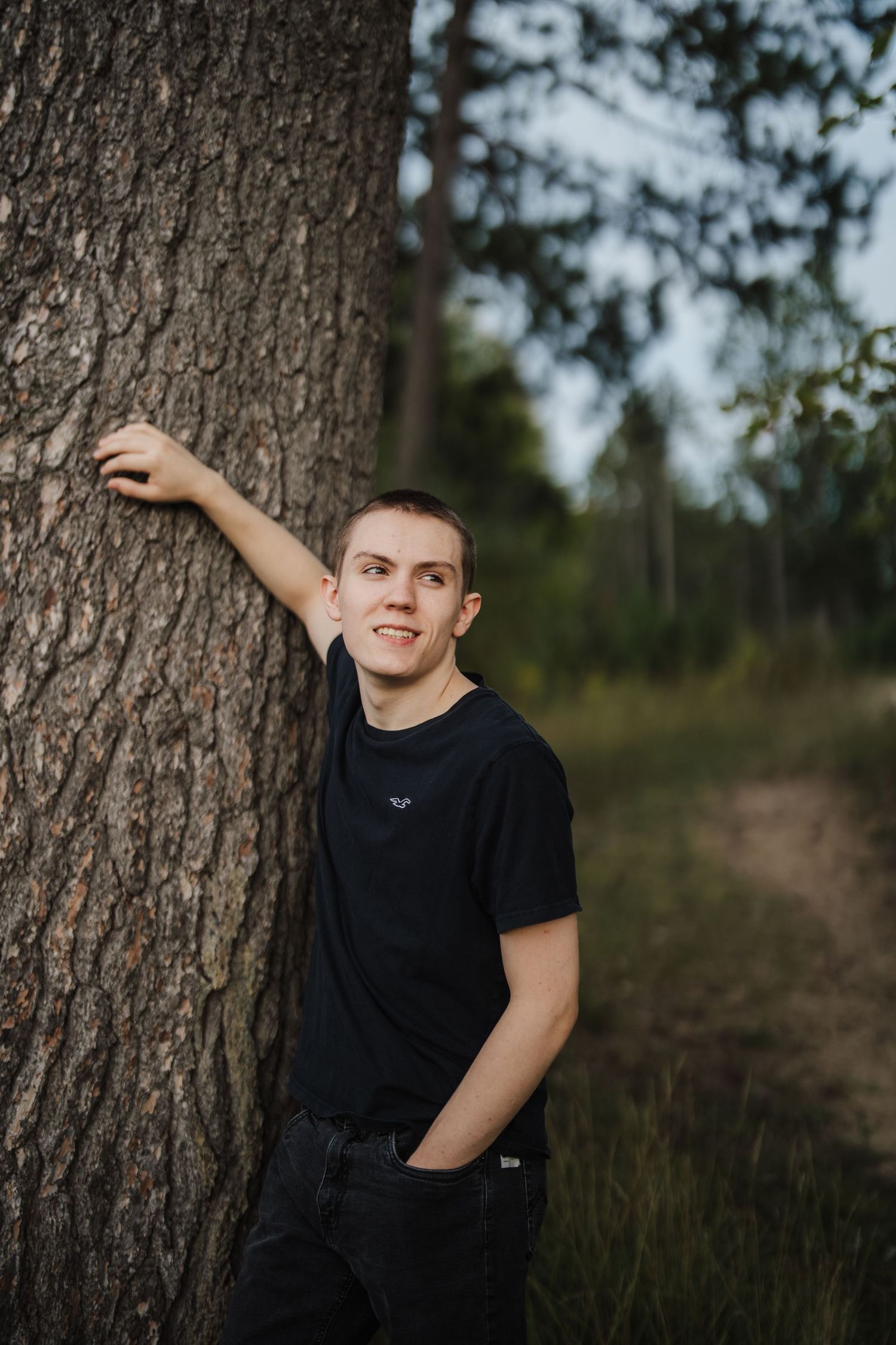 Outdoor portrait of someone in a black t-shirt leaning against a large tree trunk in a natural setting.