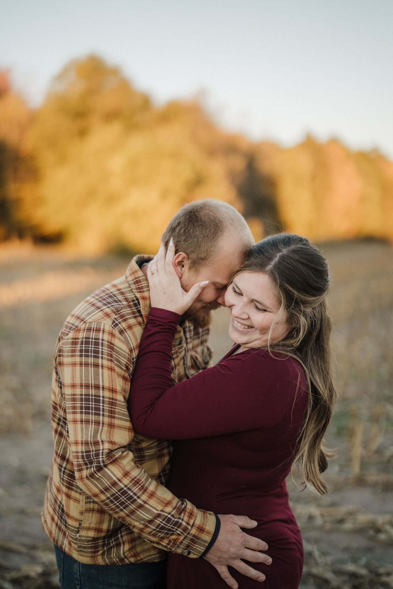 A couple embraces tenderly in a field during golden hour, wearing burgundy and plaid clothing.