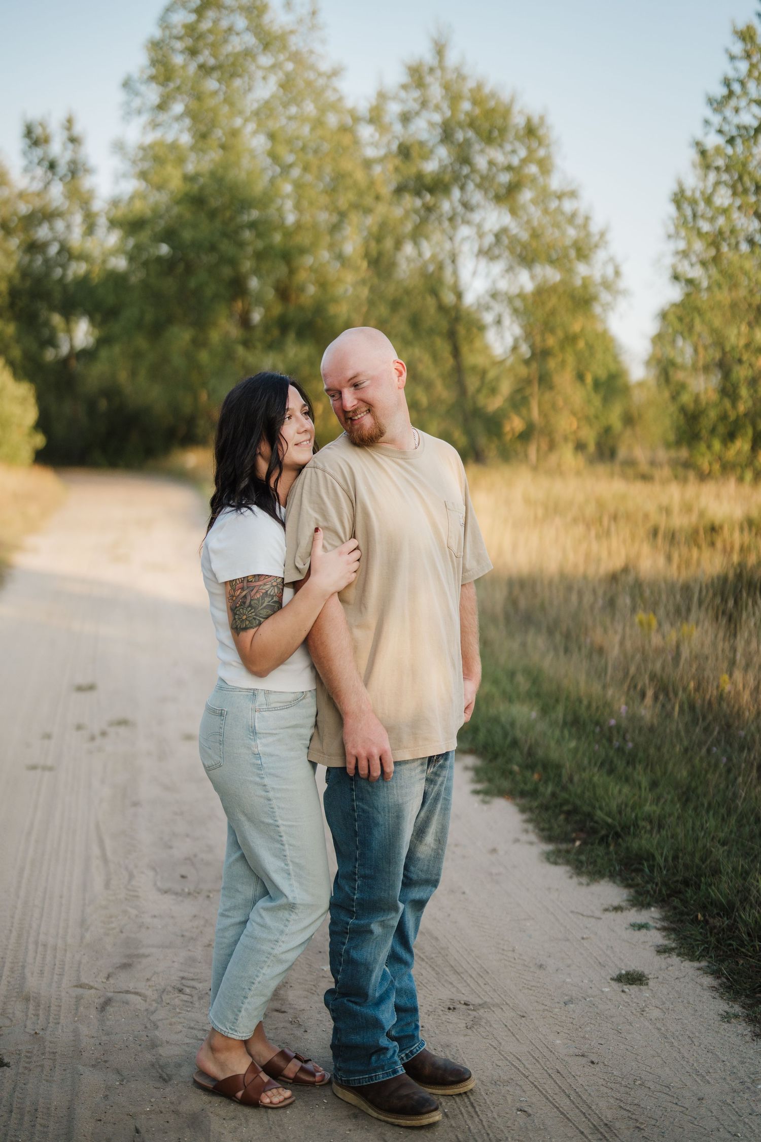 A couple shares a playful moment together on a dirt path surrounded by tall grass and trees at sunset.