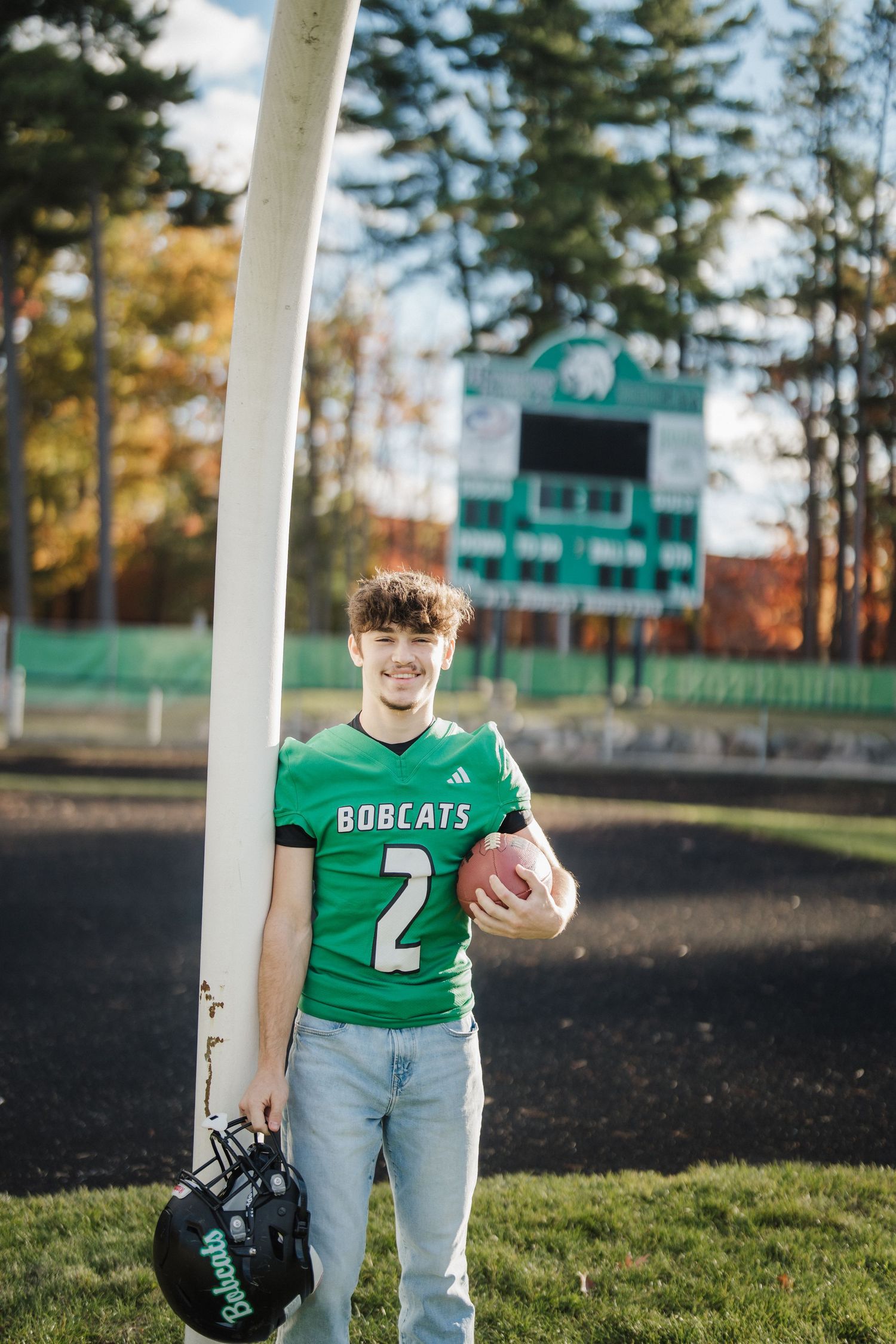 Football player in green Bobcats jersey number 2 poses with ball and helmet on field with scoreboard in background.
