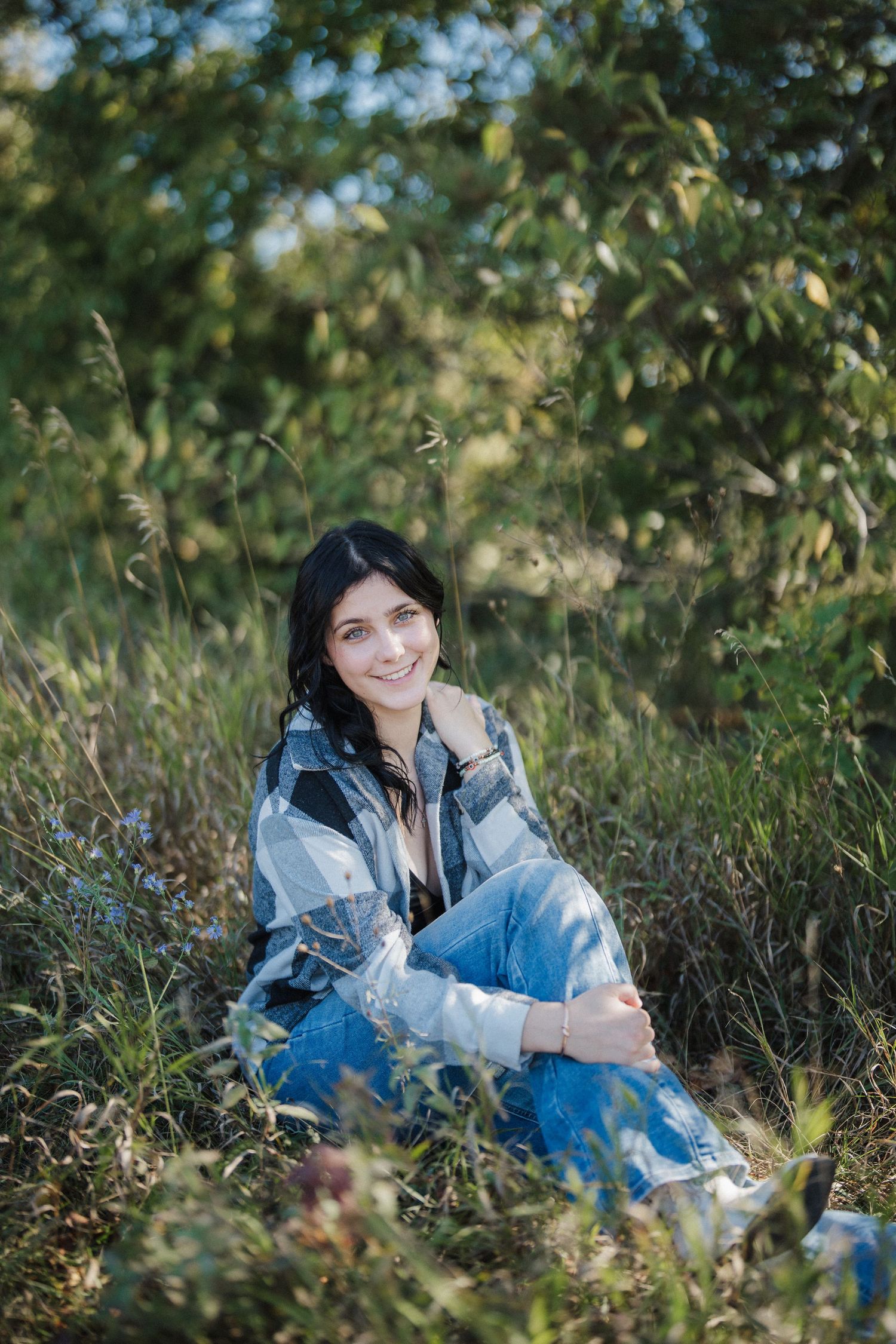 A person in denim sits casually in tall grass and greenery on a sunny day.