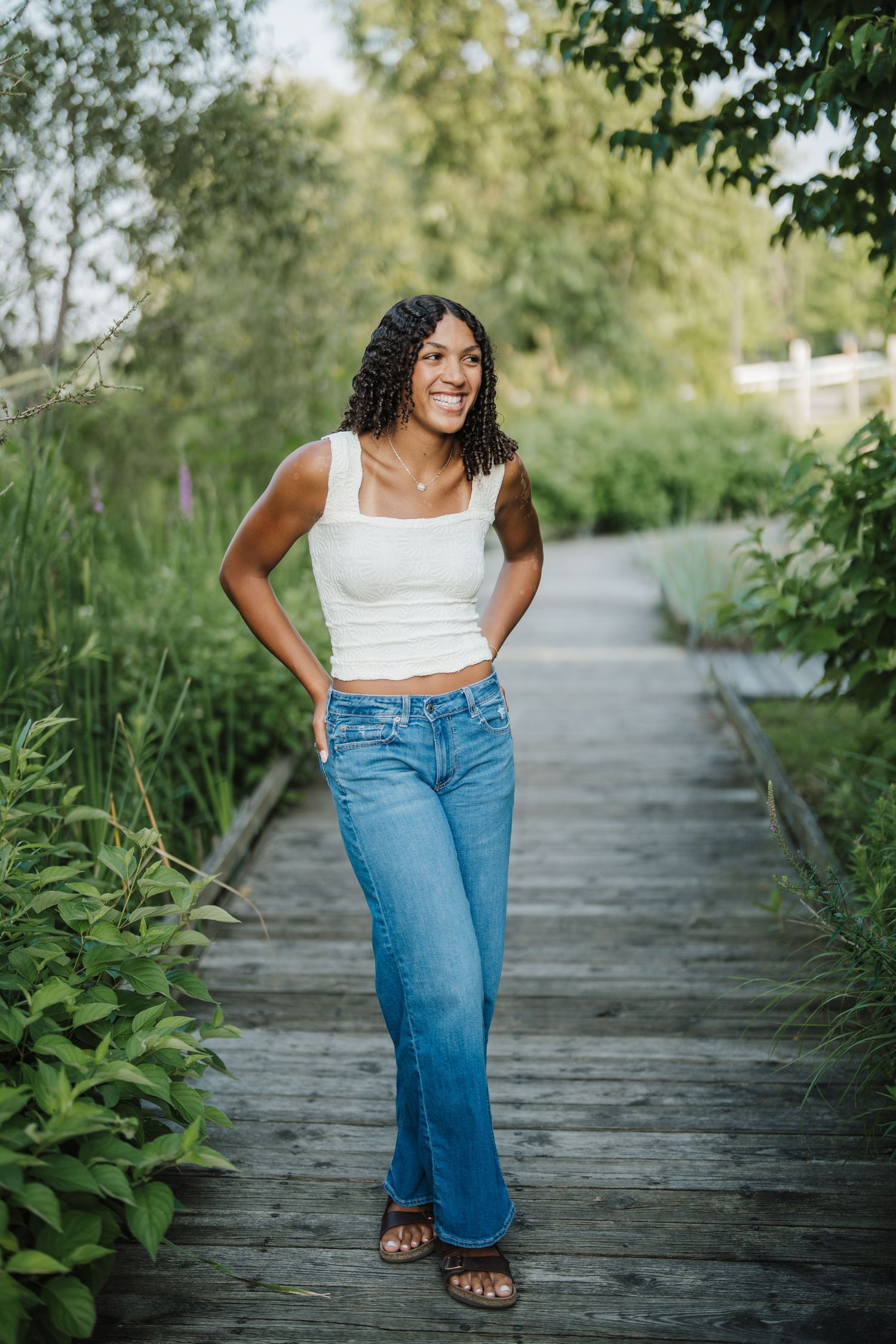 A person in blue jeans and white top walks along a wooden boardwalk surrounded by lush greenery.