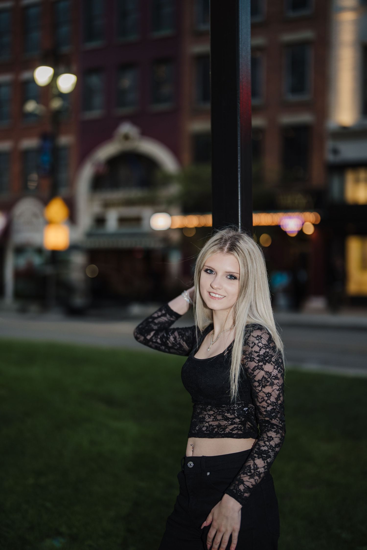A woman in a black lace crop top poses outdoors at dusk against an urban backdrop with brick buildings.