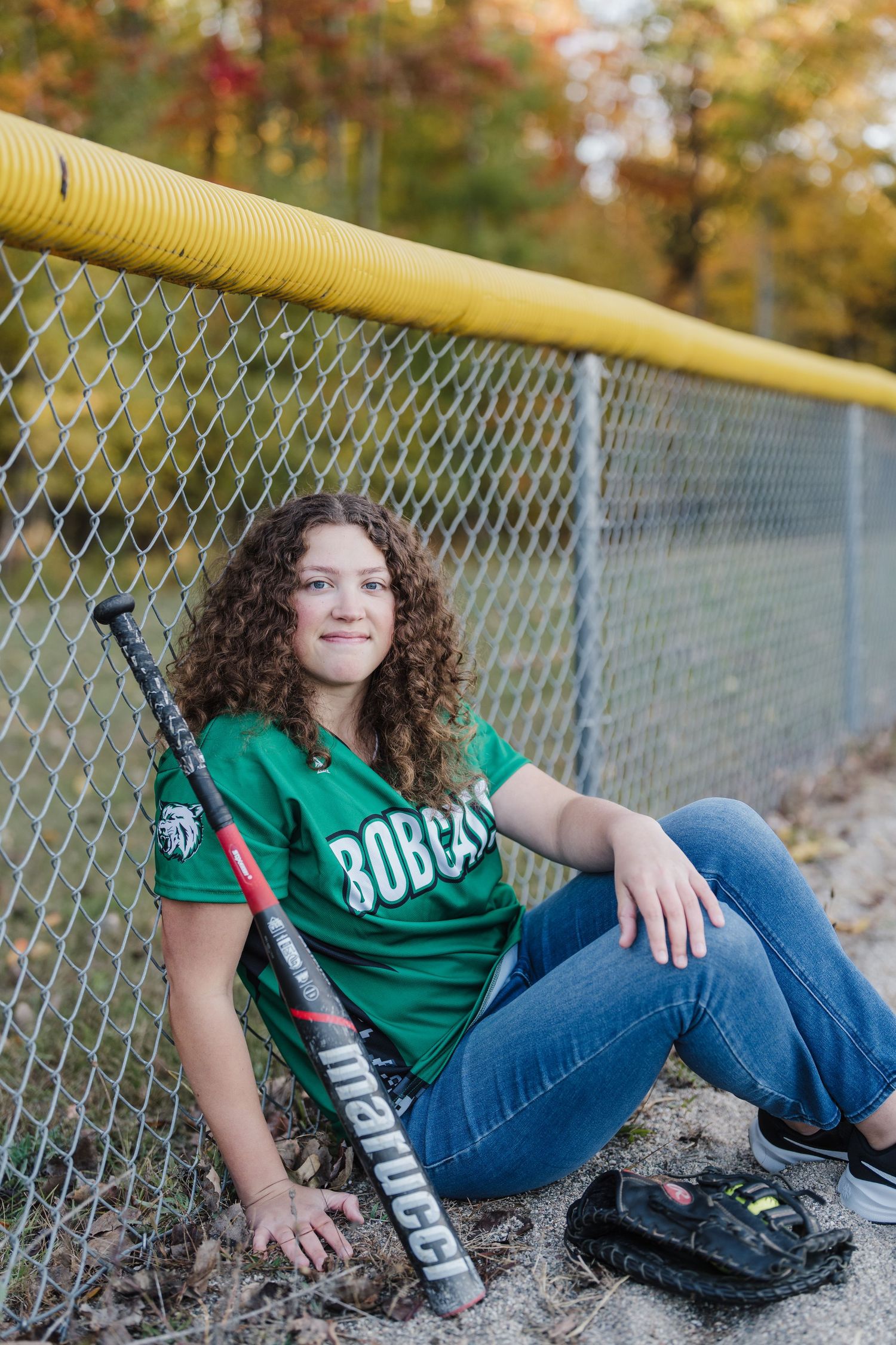 Person in green jersey and jeans lounges against yellow railing with chain link fence in autumn setting.