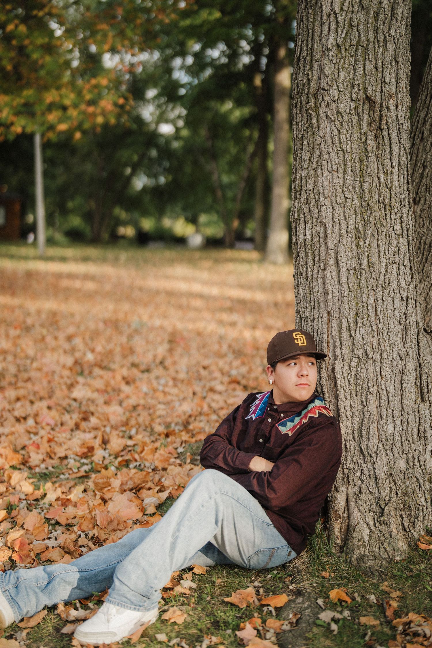 Someone relaxes against a tree trunk surrounded by fallen autumn leaves in a peaceful park setting.