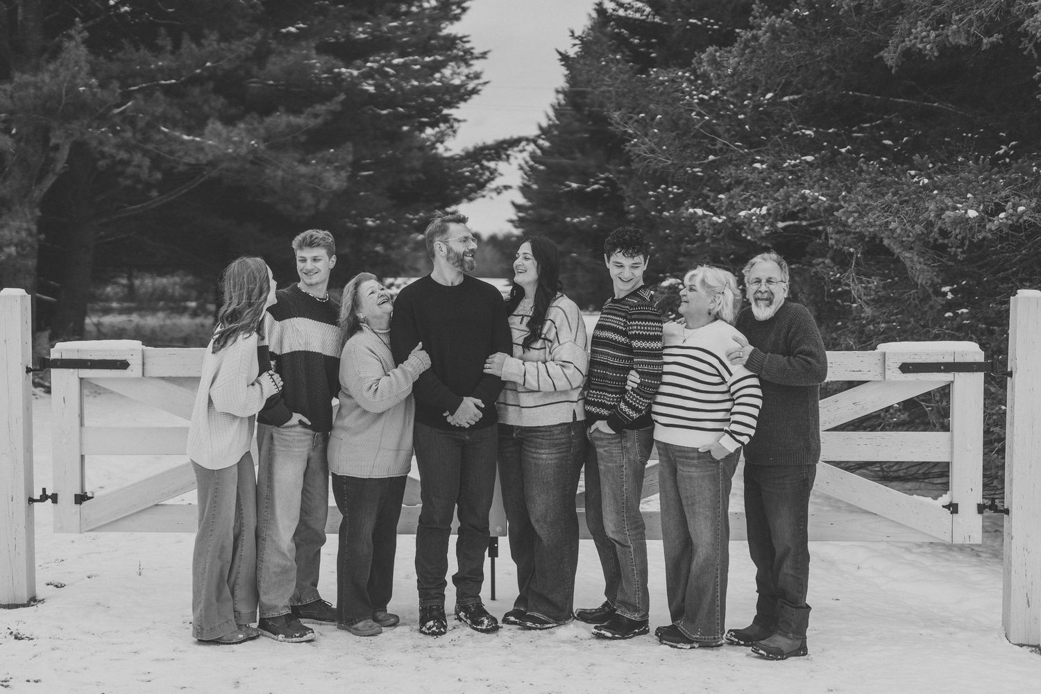A group of people in casual winter clothing stand together by a white gate on a tree-lined path in black and white.