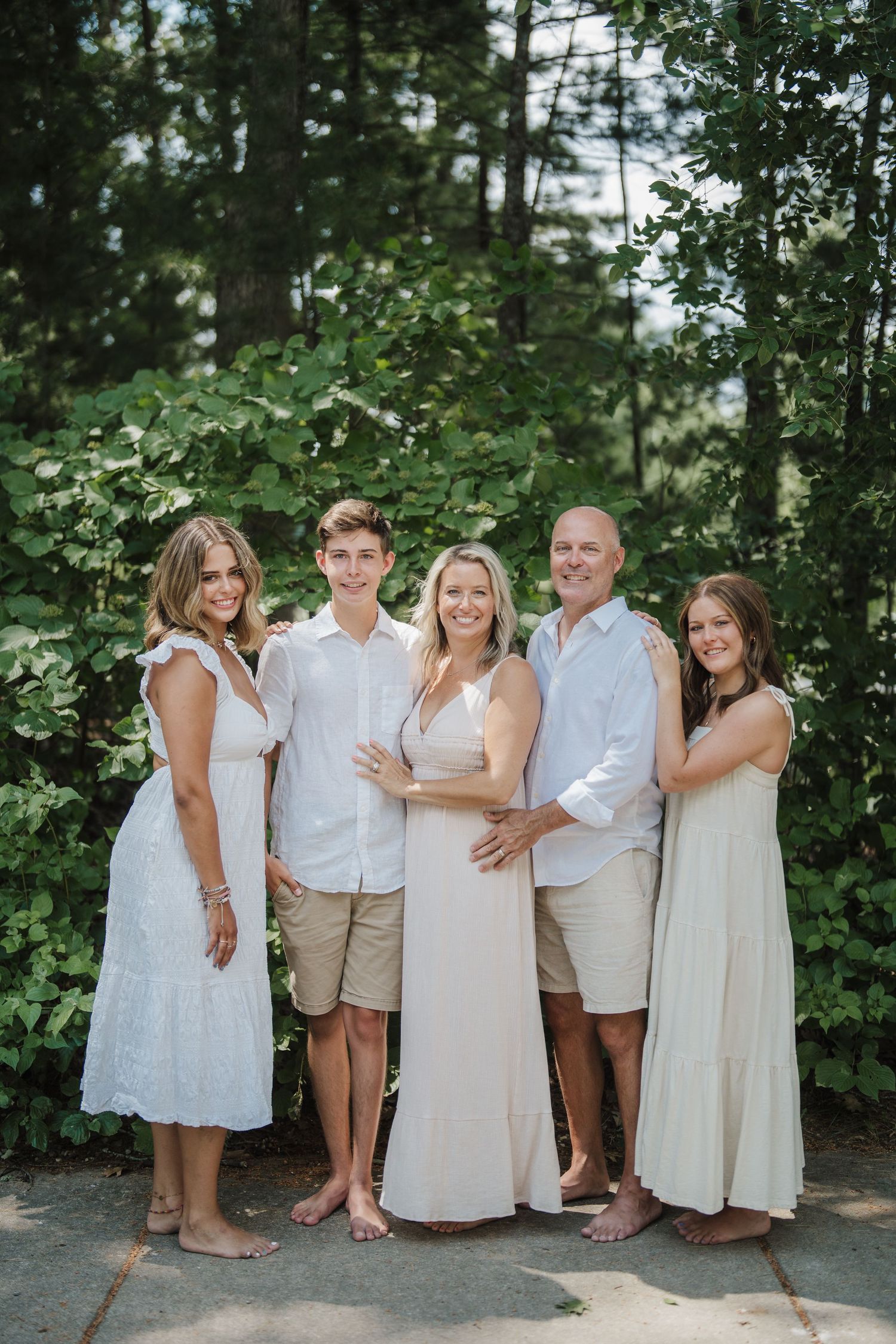 A family of five poses together outdoors wearing coordinated white and beige outfits against a lush green backdrop.