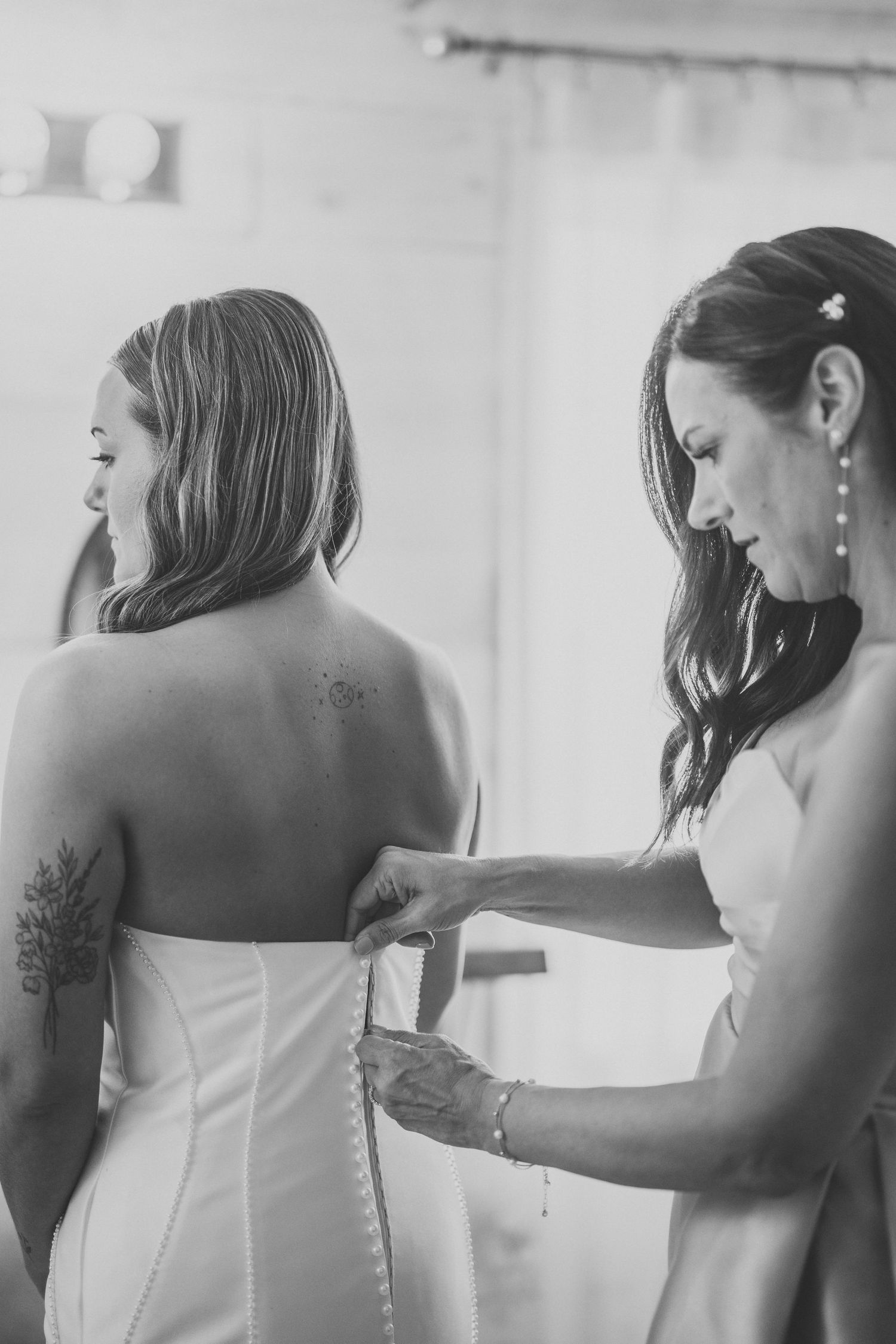 Black and white photo of a bride getting help with her dress buttons during wedding preparations.