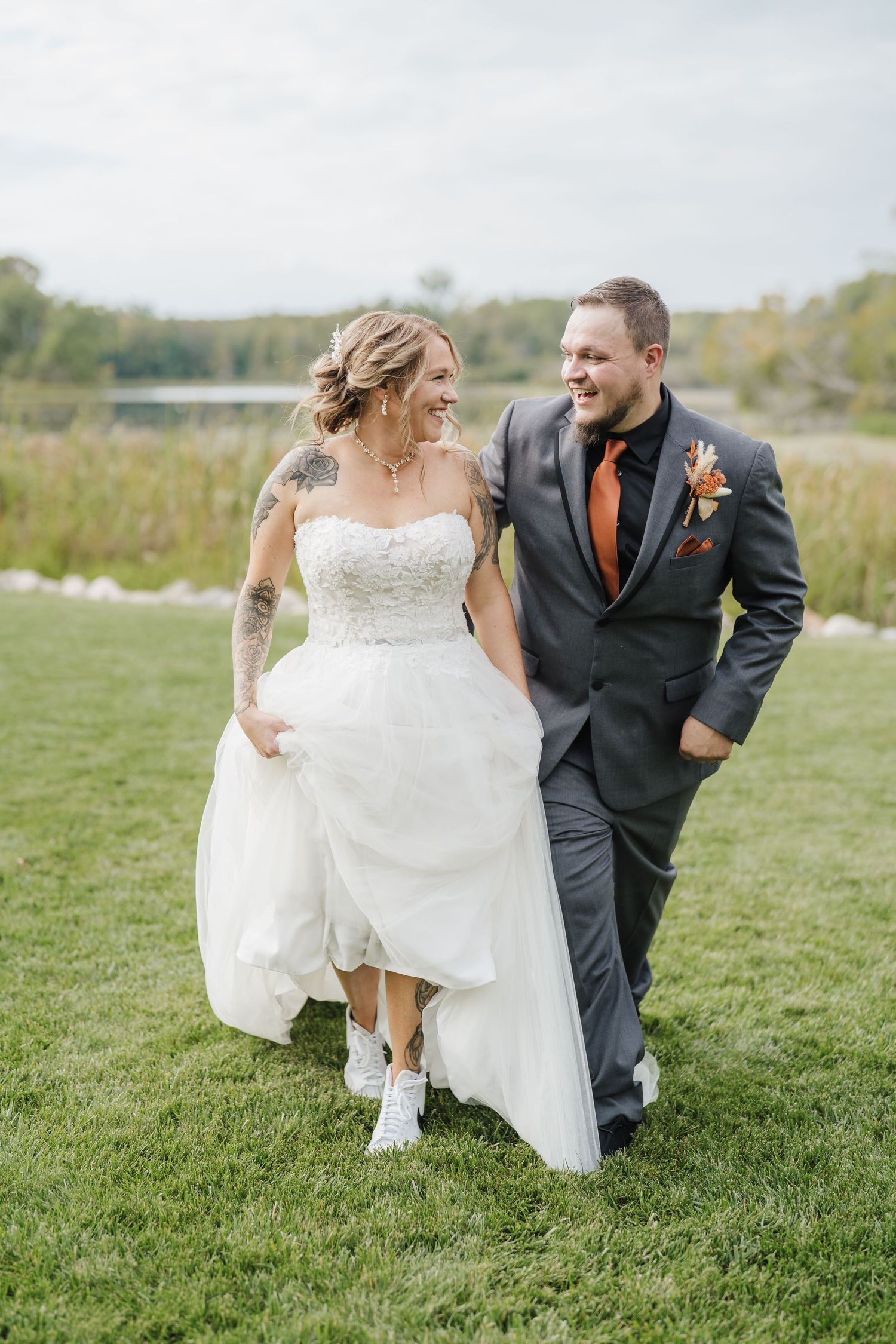 A couple in wedding attire playfully runs across a grassy field on their special day.