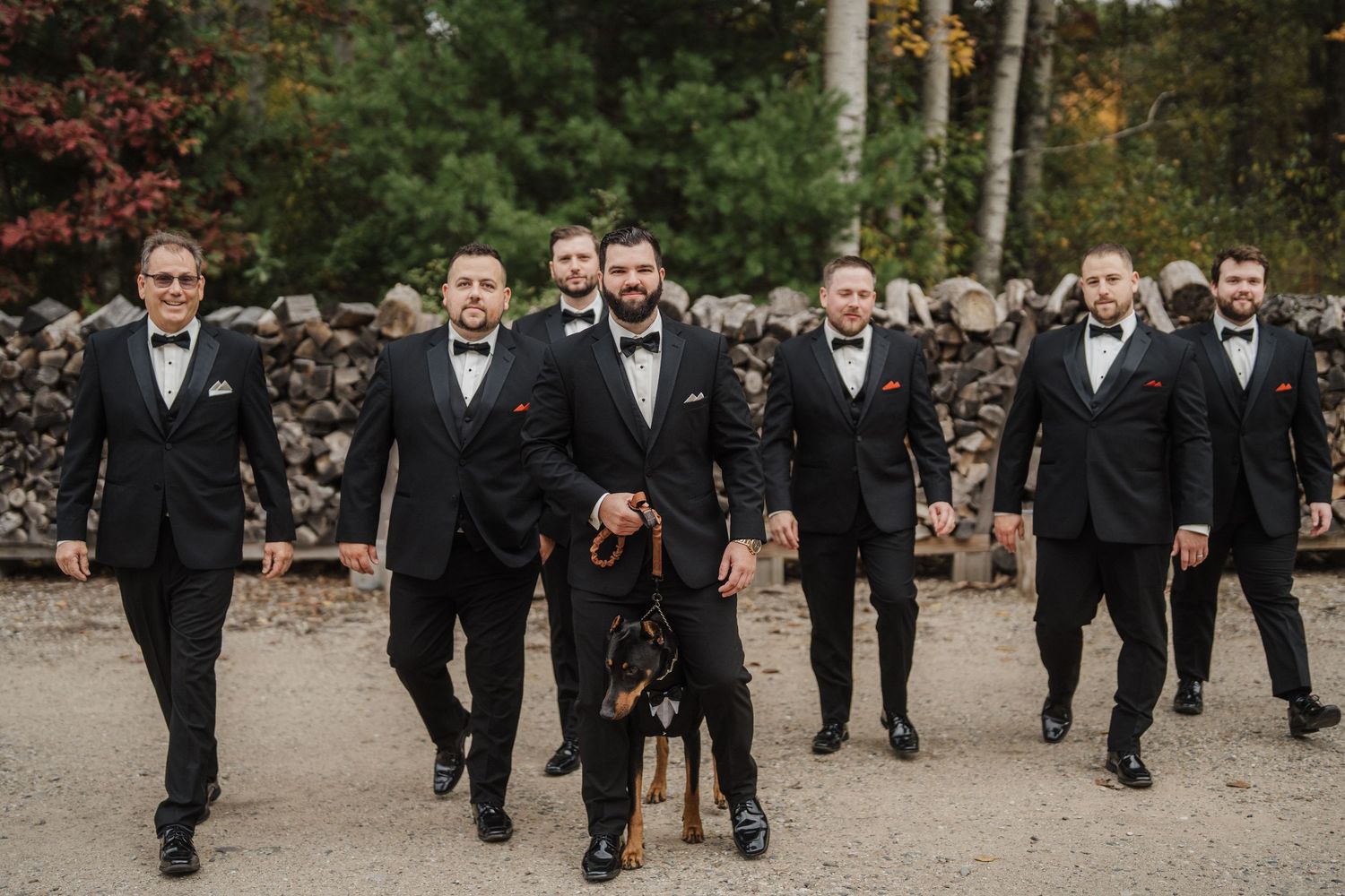 Groomsmen in black tuxedos walk together with a black dog on a path during a wedding celebration.