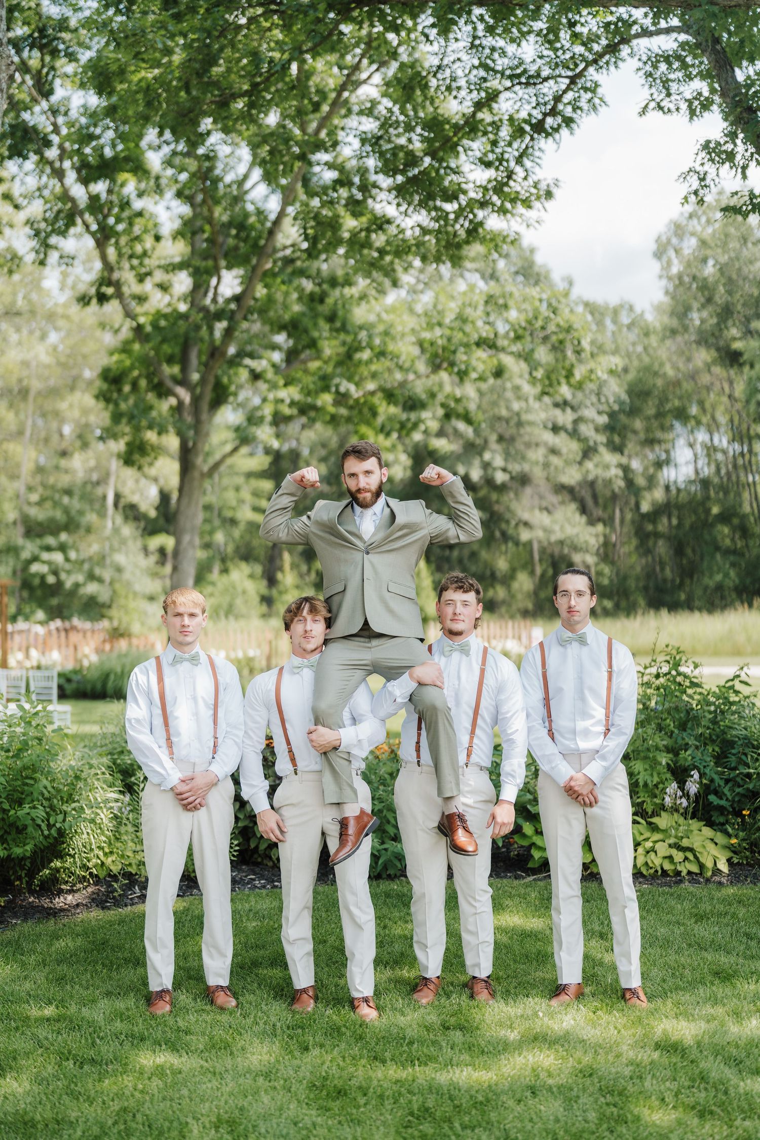 Groomsmen in white suspenders pose while one jumps in the air against a garden backdrop.