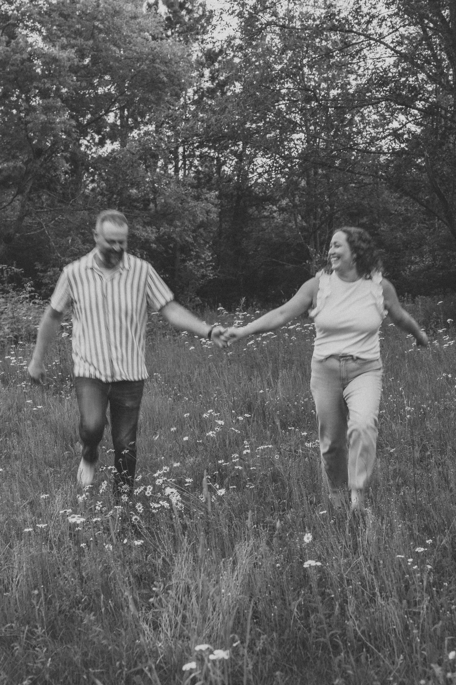 Black and white photo of a happy couple holding hands and running through a wildflower meadow with trees behind them.