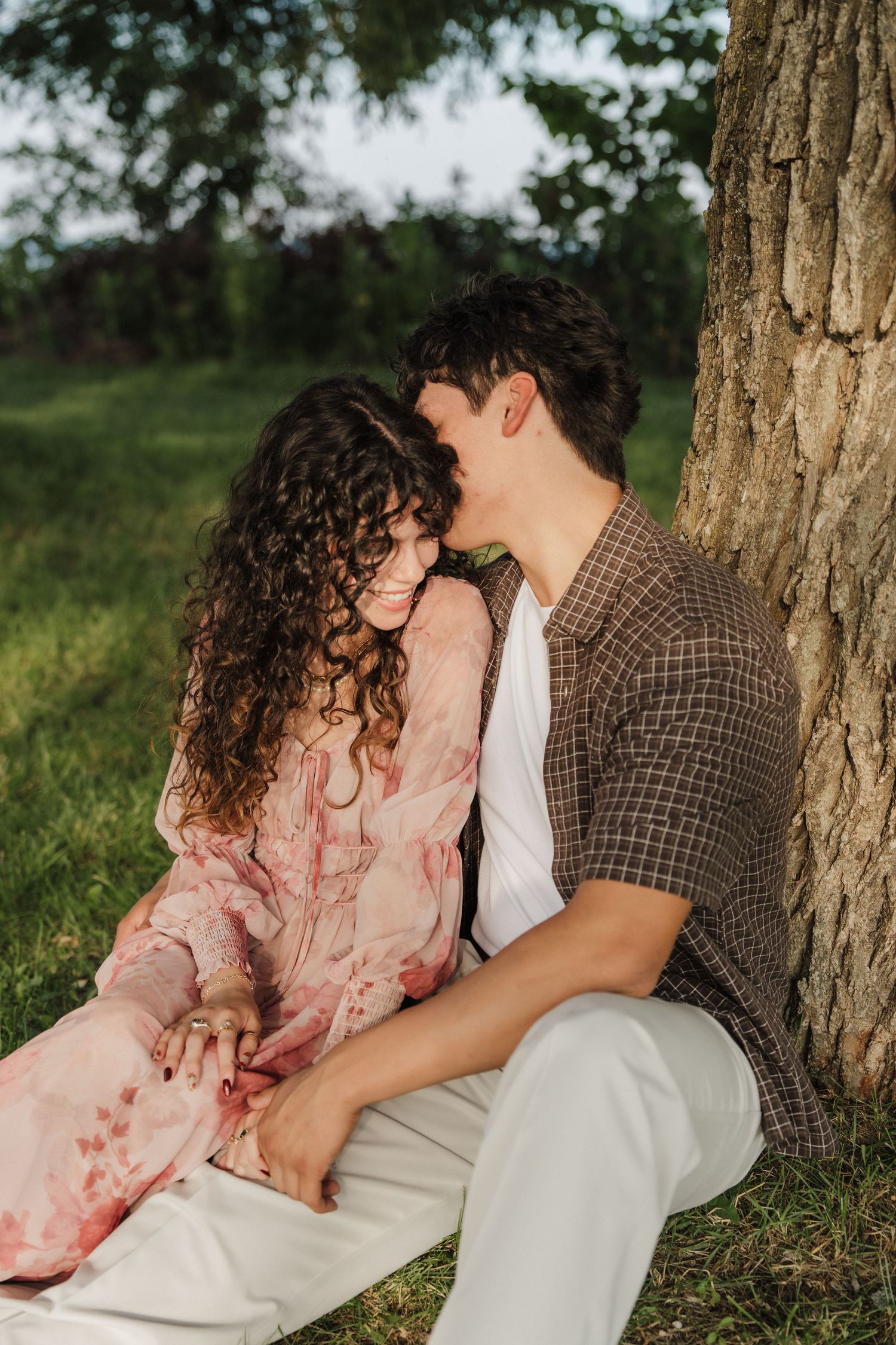 A couple sits together leaning against a tree in a lush green park, sharing a tender romantic moment.